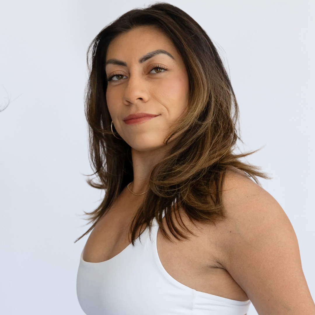 A woman with long wavy brown hair smiling and wearing a black cropped hoodie that says 'REBEL HOUSE' in white text, standing in front of a plain white wall.