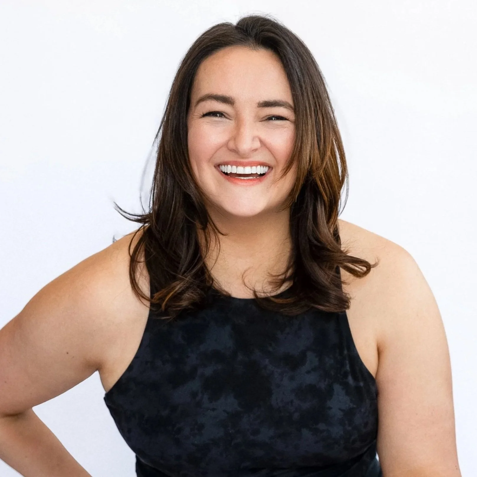 A woman with shoulder-length dark brown hair, smiling, wearing a black sleeveless top, standing against a white background.