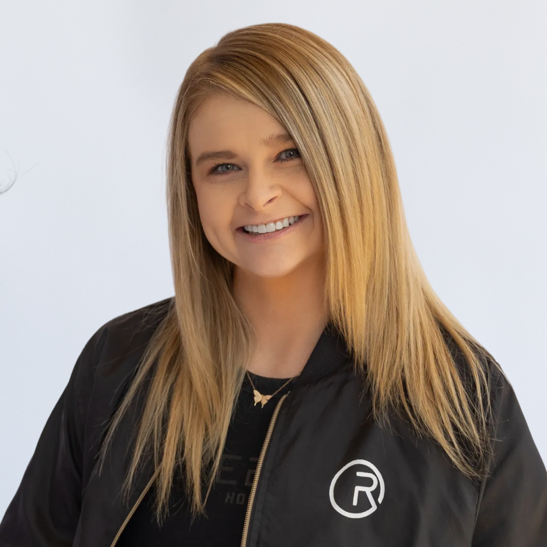 A smiling woman with straight, shoulder-length blonde hair wearing a black t-shirt with a colorful circular logo on the front.