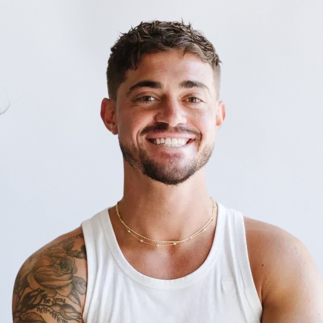 Smiling young man with tattoos on his left arm, wearing a white sleeveless shirt and a fitness glove on his right wrist, standing against a plain white background.