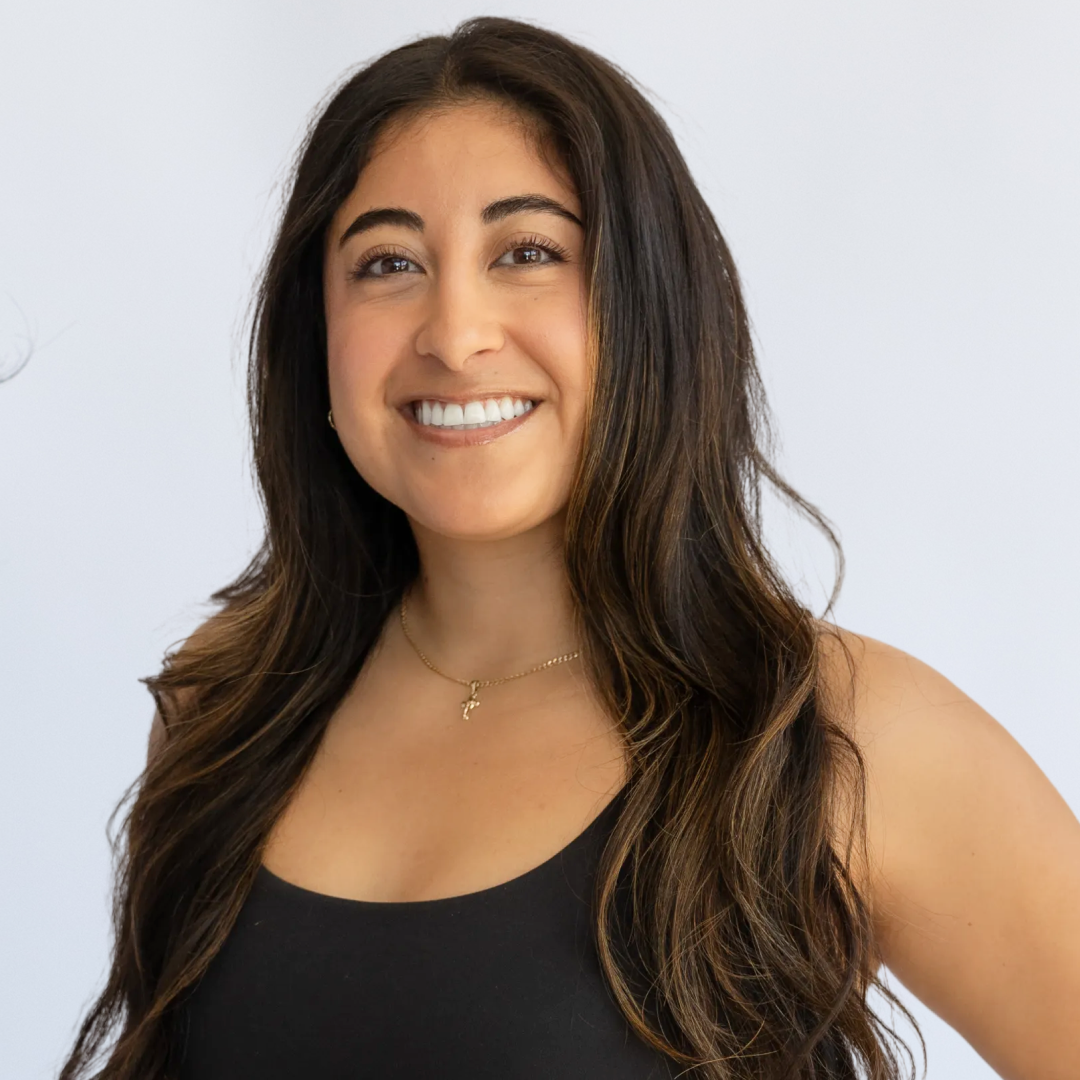 A woman with long brown hair, wearing a black crop top and black pants, smiling at the camera with her hand on her hip, against a plain white background.