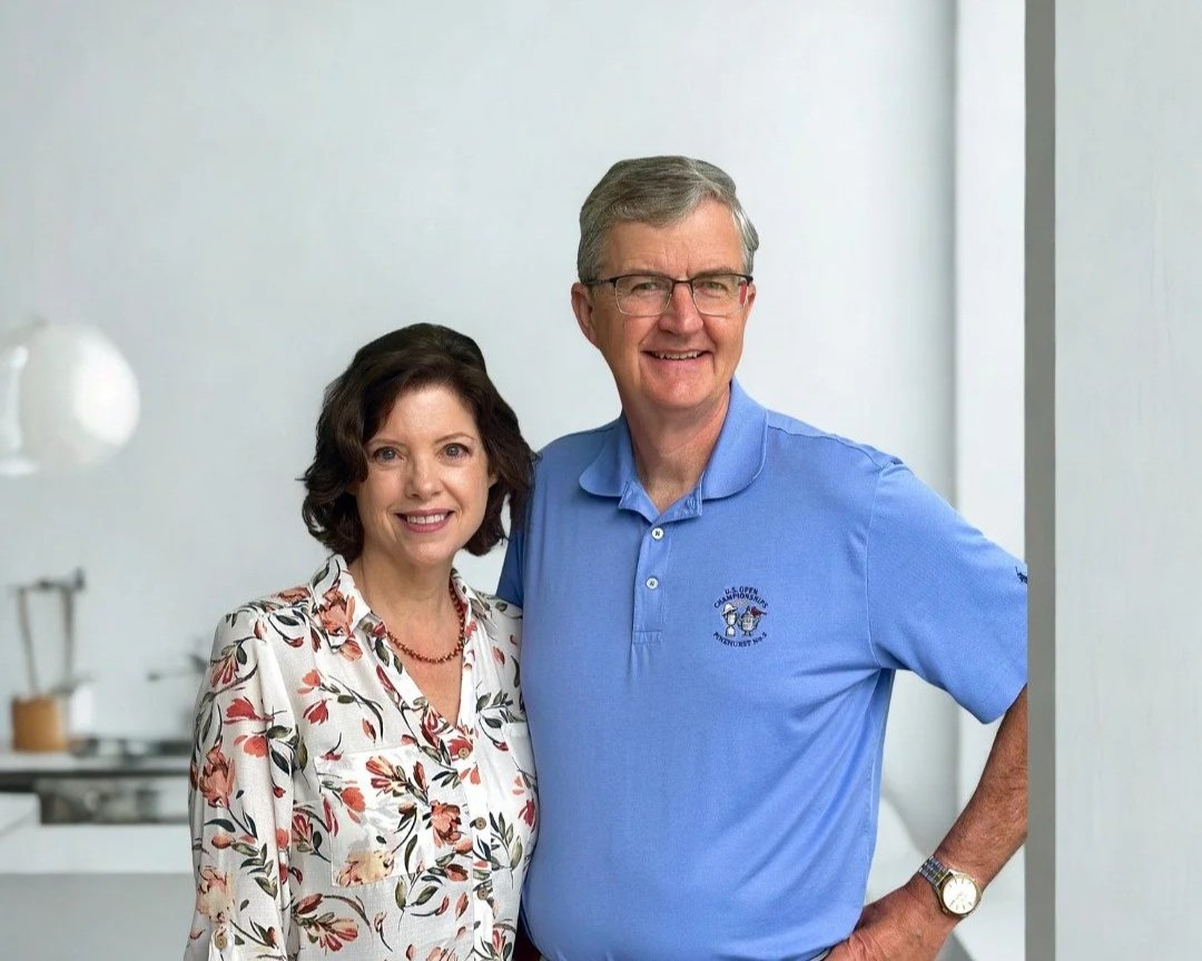 A smiling woman with dark wavy hair wearing a floral blouse and a man with glasses and gray hair wearing a blue polo shirt, standing together in front of a pink and gray background.