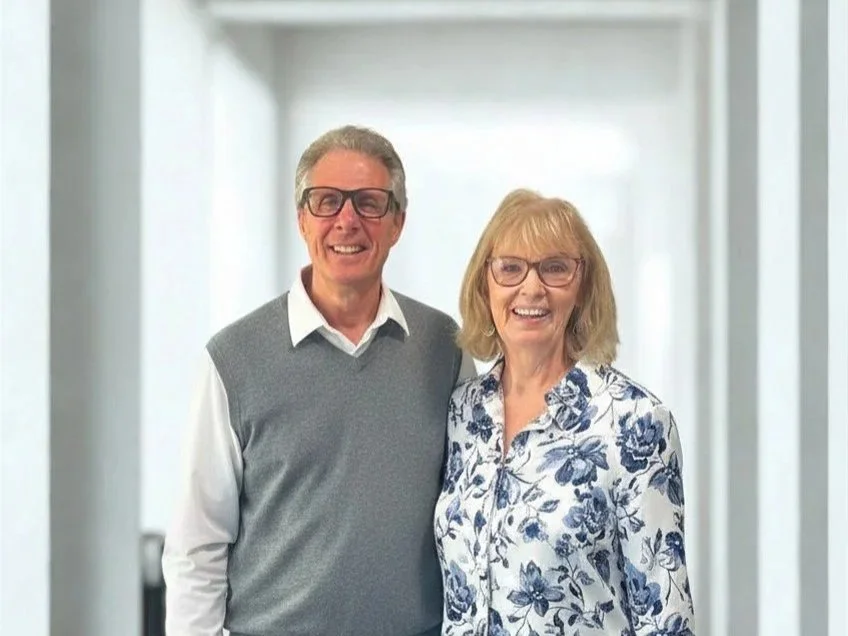Smiling older man and woman standing together against a red circular backdrop, both wearing glasses and casual clothing.