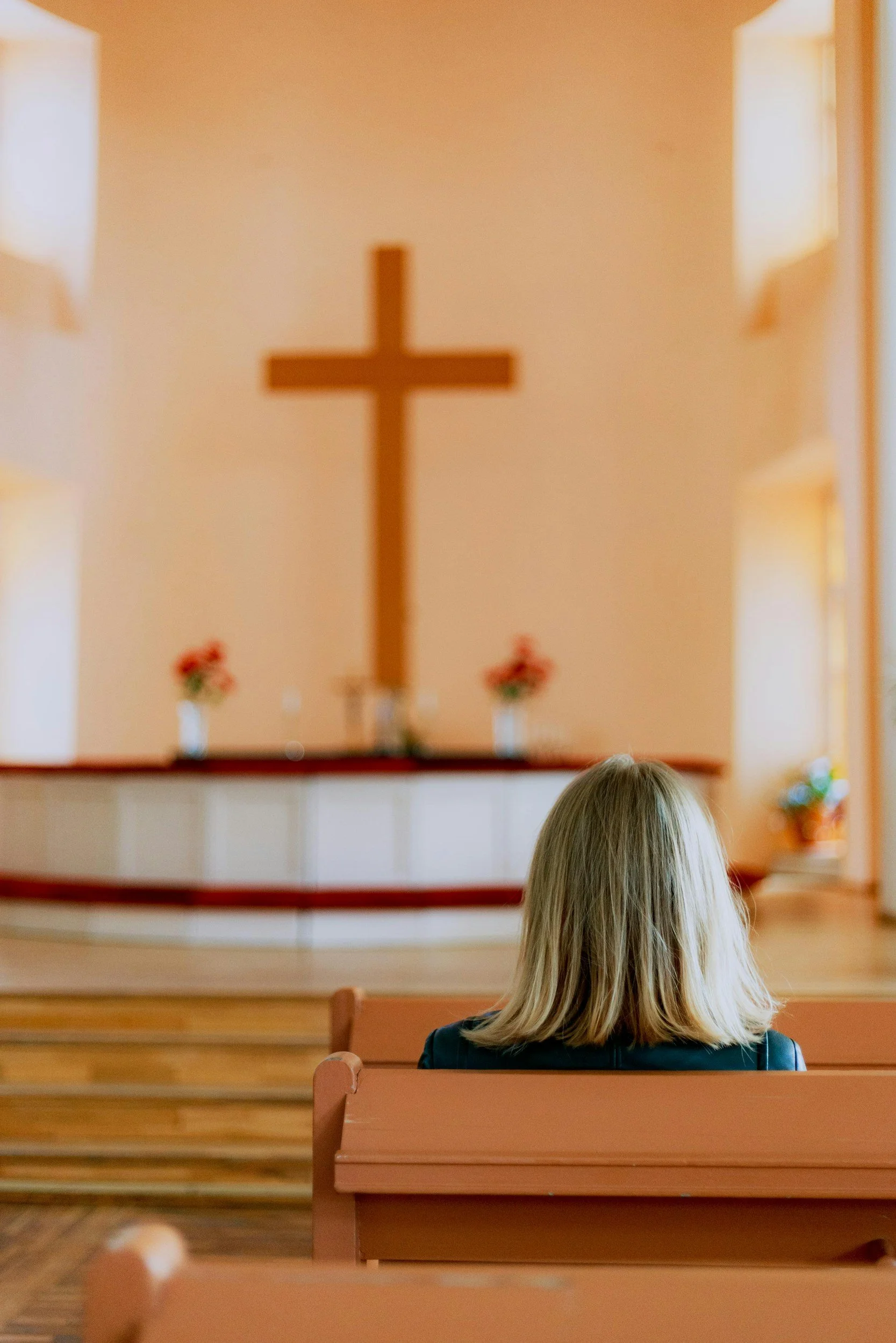 A woman sitting on a wooden pew inside a church, facing an altar with a large cross and flower arrangements.