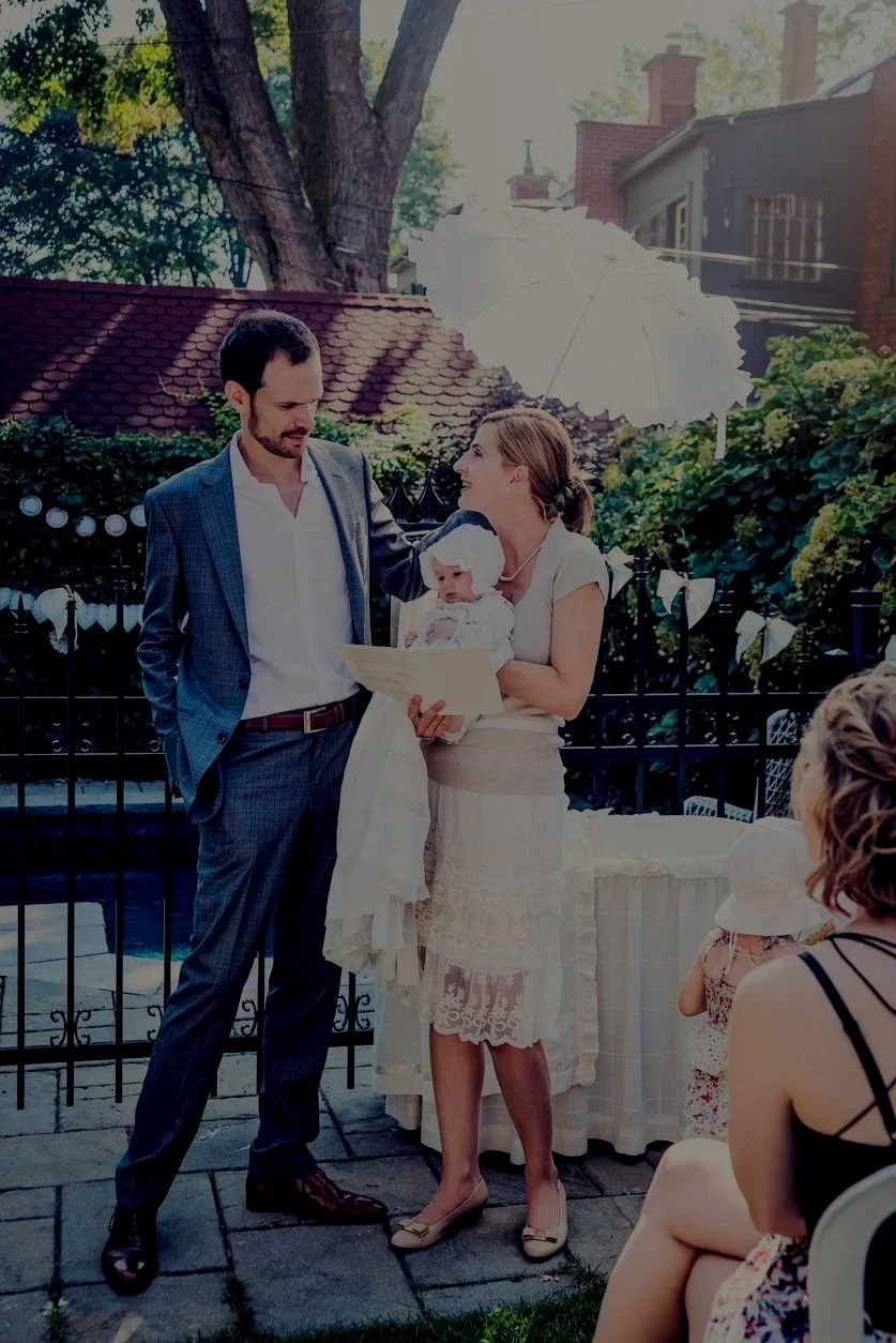 A couple standing outdoors at a wedding or celebration, with a woman holding a baby, a white parasol overhead, and others seated nearby.