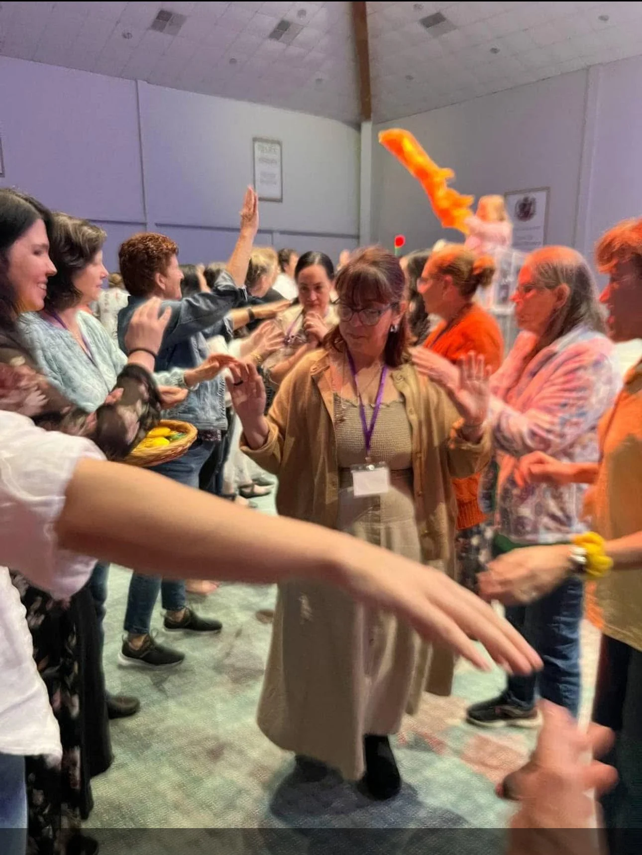 A group of women standing in lines facing each other at an indoor event, some raising their hands, with one woman in the center wearing a beige coat and glasses, and a woman in the background. Ladies meeting.