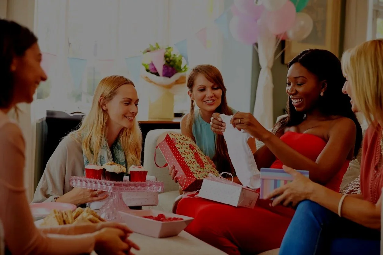 A group of five women gathered in a living room celebrating a birthday, with balloons, present boxes, cupcakes, and snacks on the table.