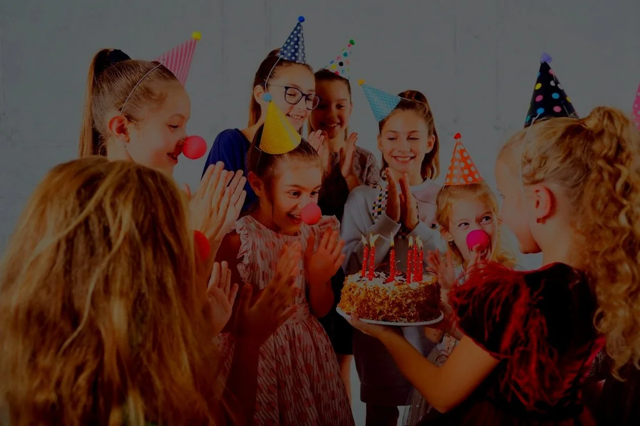 A group of children celebrating a birthday, wearing party hats and clown noses, with a girl holding a birthday cake with lit candles.
