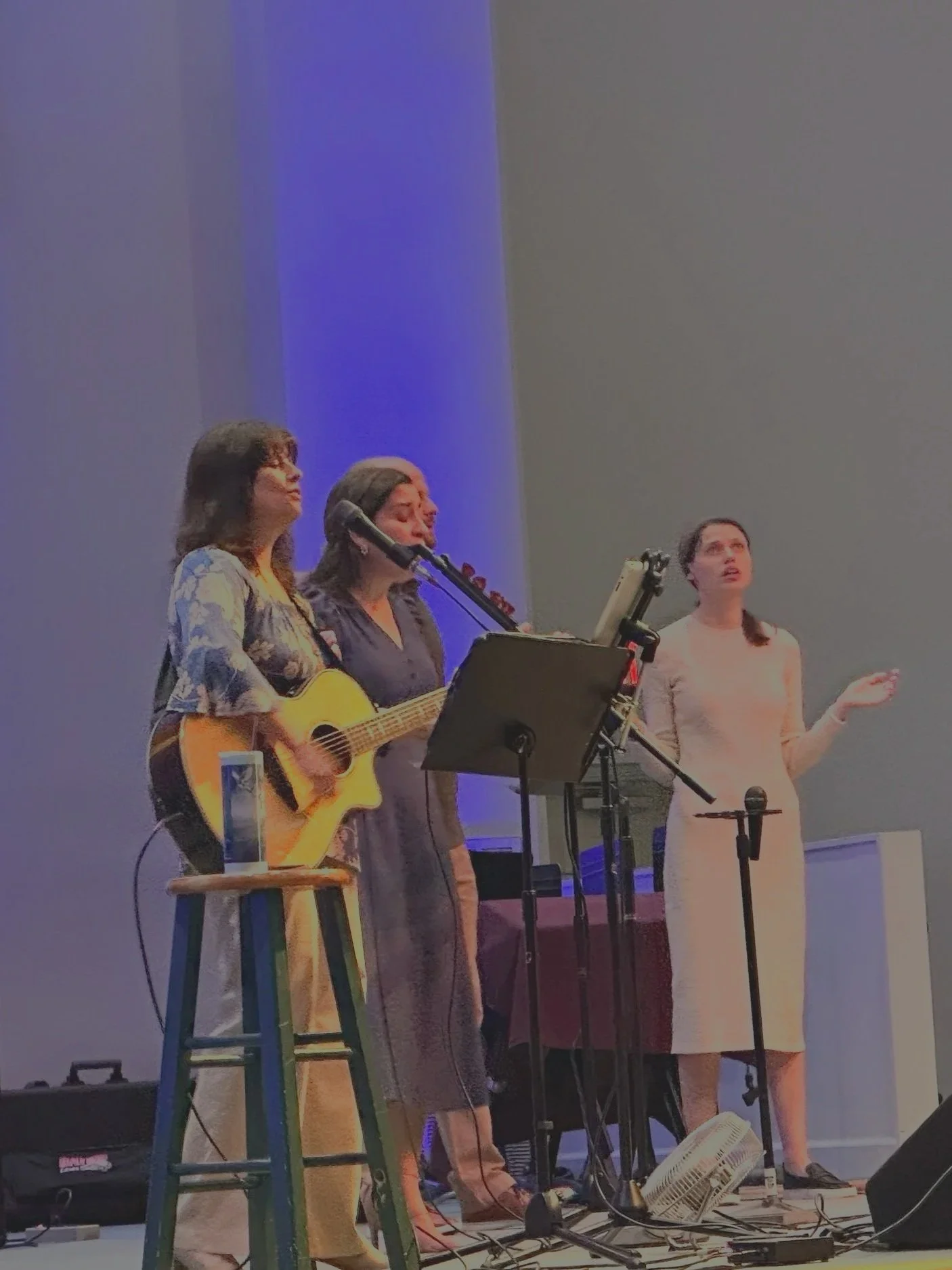 Three women performing on stage, one playing guitar, one singing into a microphone, and one gesturing with her hand, with microphone stands and music stands around them. Worship team