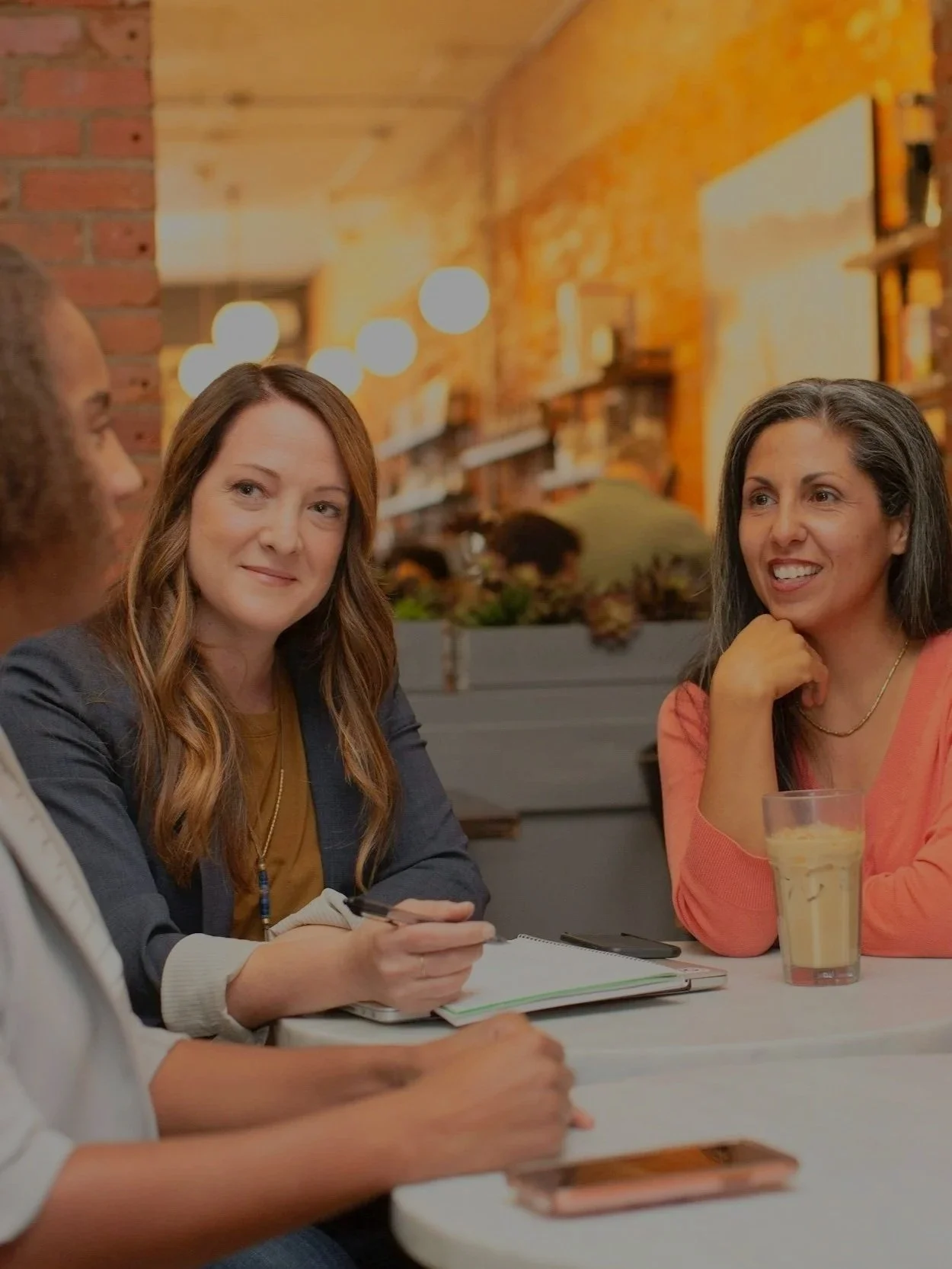 Four women sitting at a table in a cozy, warmly lit cafe or restaurant, engaged in conversation. One woman has a notepad and pen, another has a drink with a straw, and they appear to be having an animated discussion.