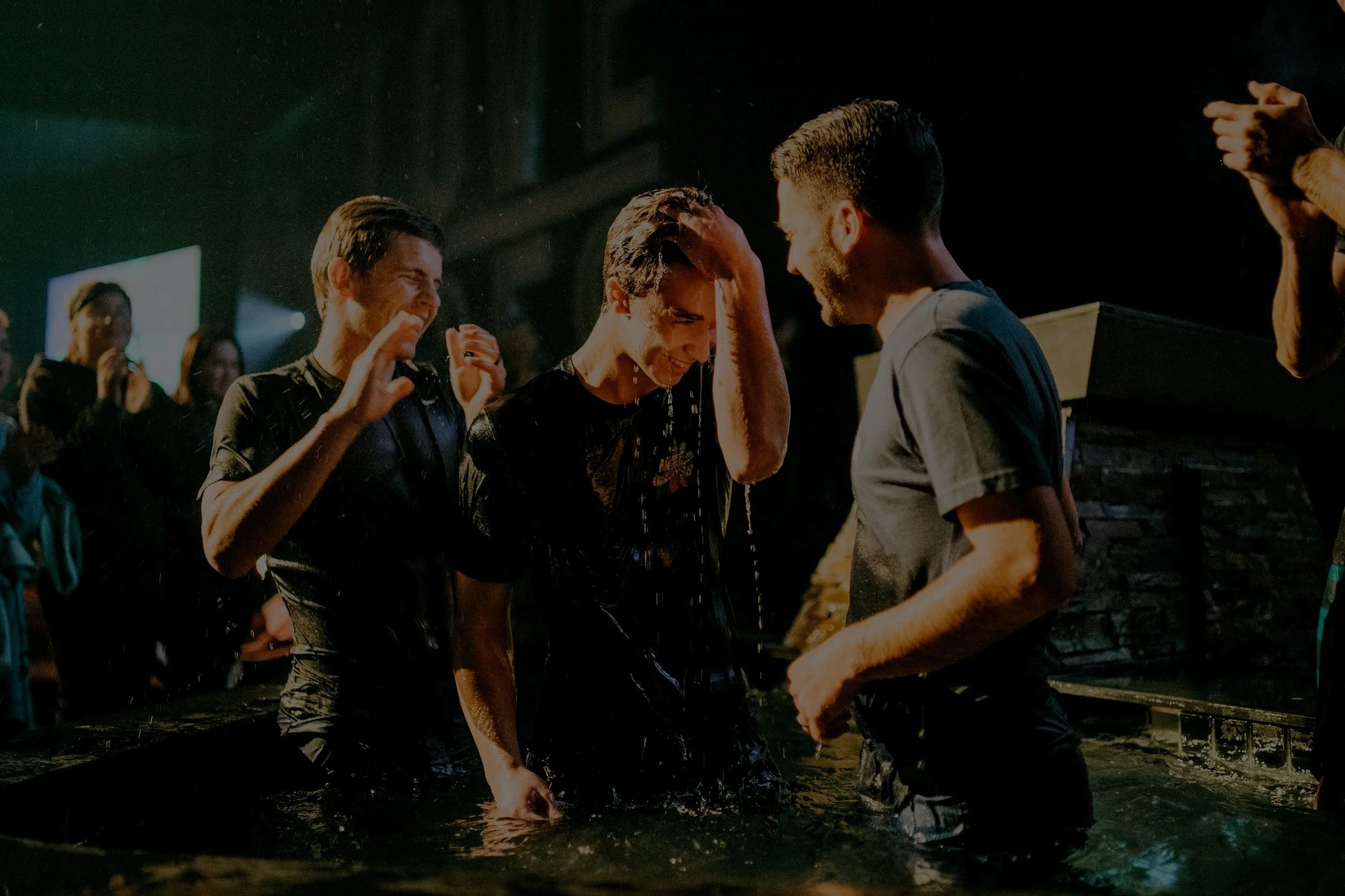 People being baptized in water baptism ceremony at night, with some smiling and others in prayer, surrounded by onlookers.