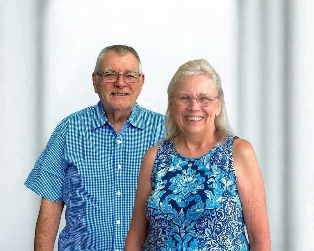 A smiling elderly couple standing in front of a gray background with a red circle behind them. The man is wearing glasses and a blue checkered shirt, and the woman is wearing glasses and a sleeveless blue floral top.