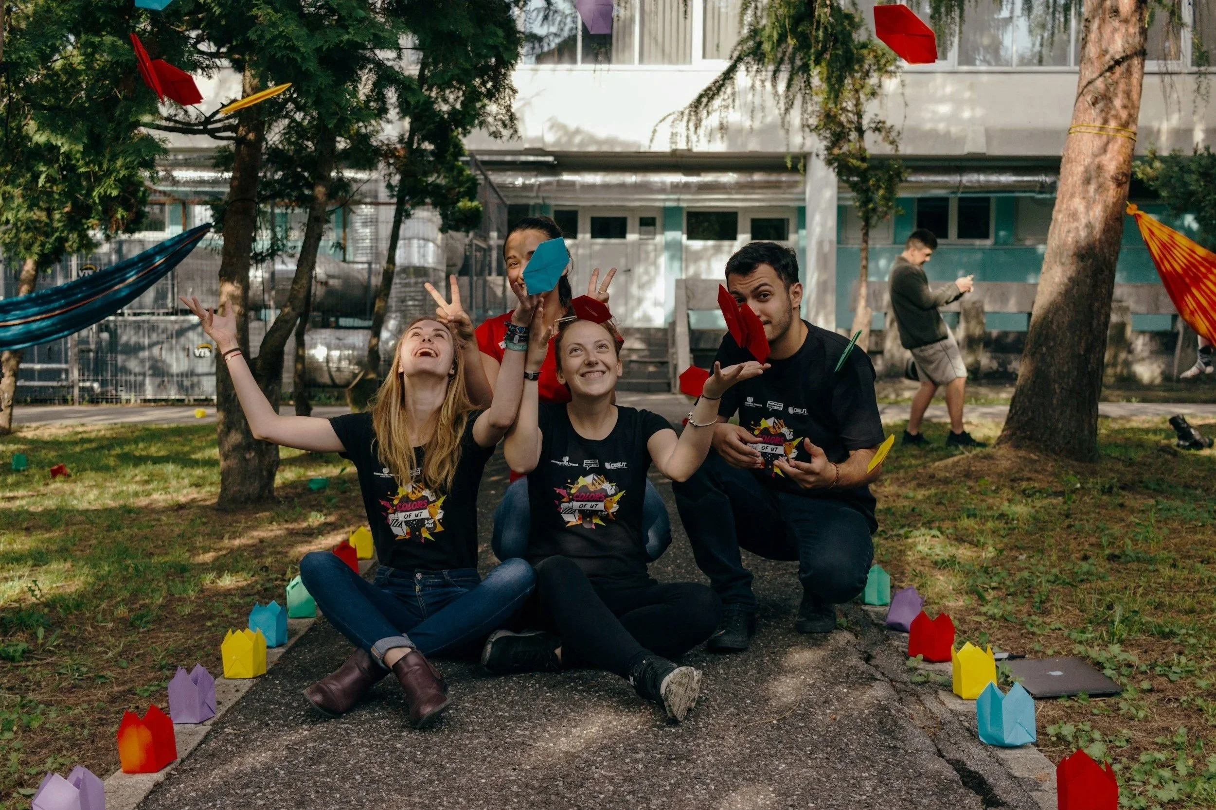 Group of kids sitting and standing on a pathway in a park, celebrating amidst colorful paper lanterns and hanging decorations, with trees and a building in the background.