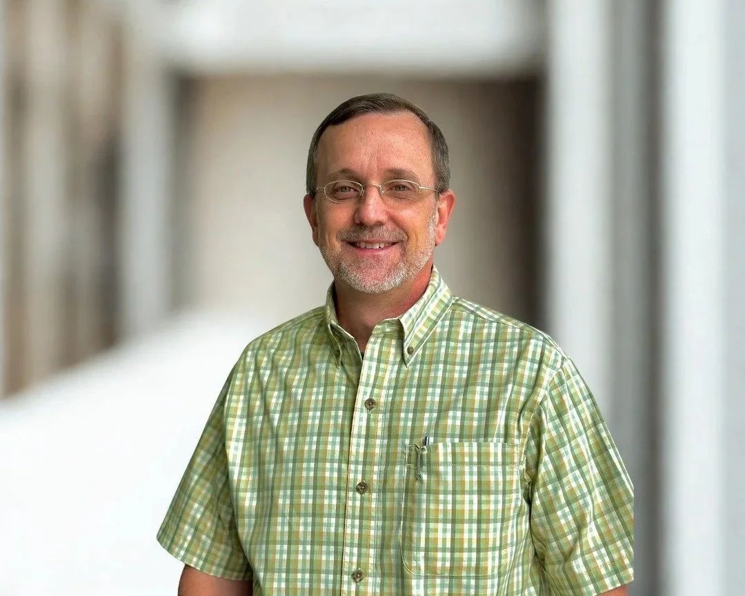 A man with glasses and short hair wearing a green plaid shirt, smiling, with a gray background and a large red circle behind him.