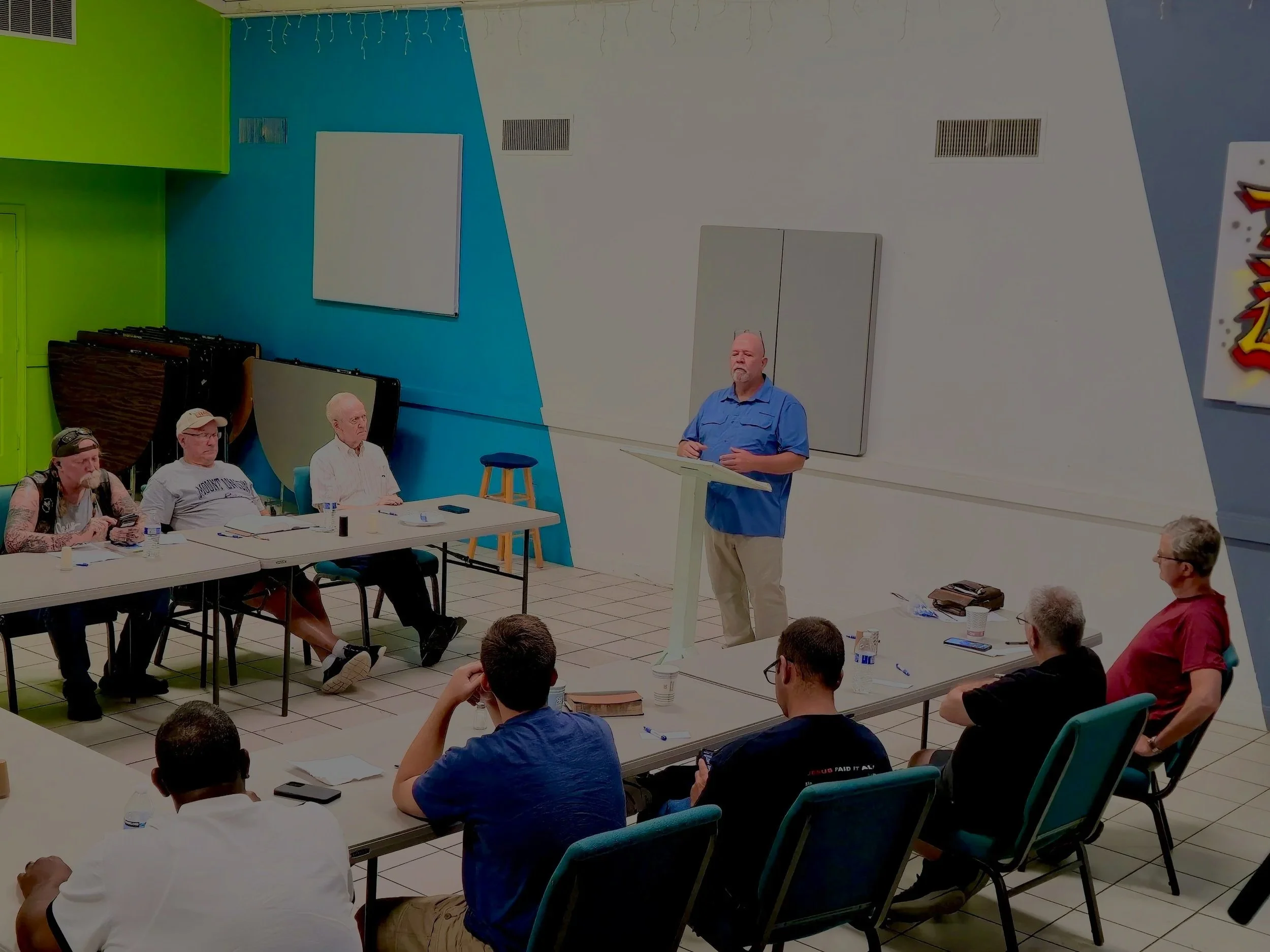 A man in a blue shirt standing at a lectern speaking to a group of people seated at tables in a brightly colored room with blue, green, and white walls.