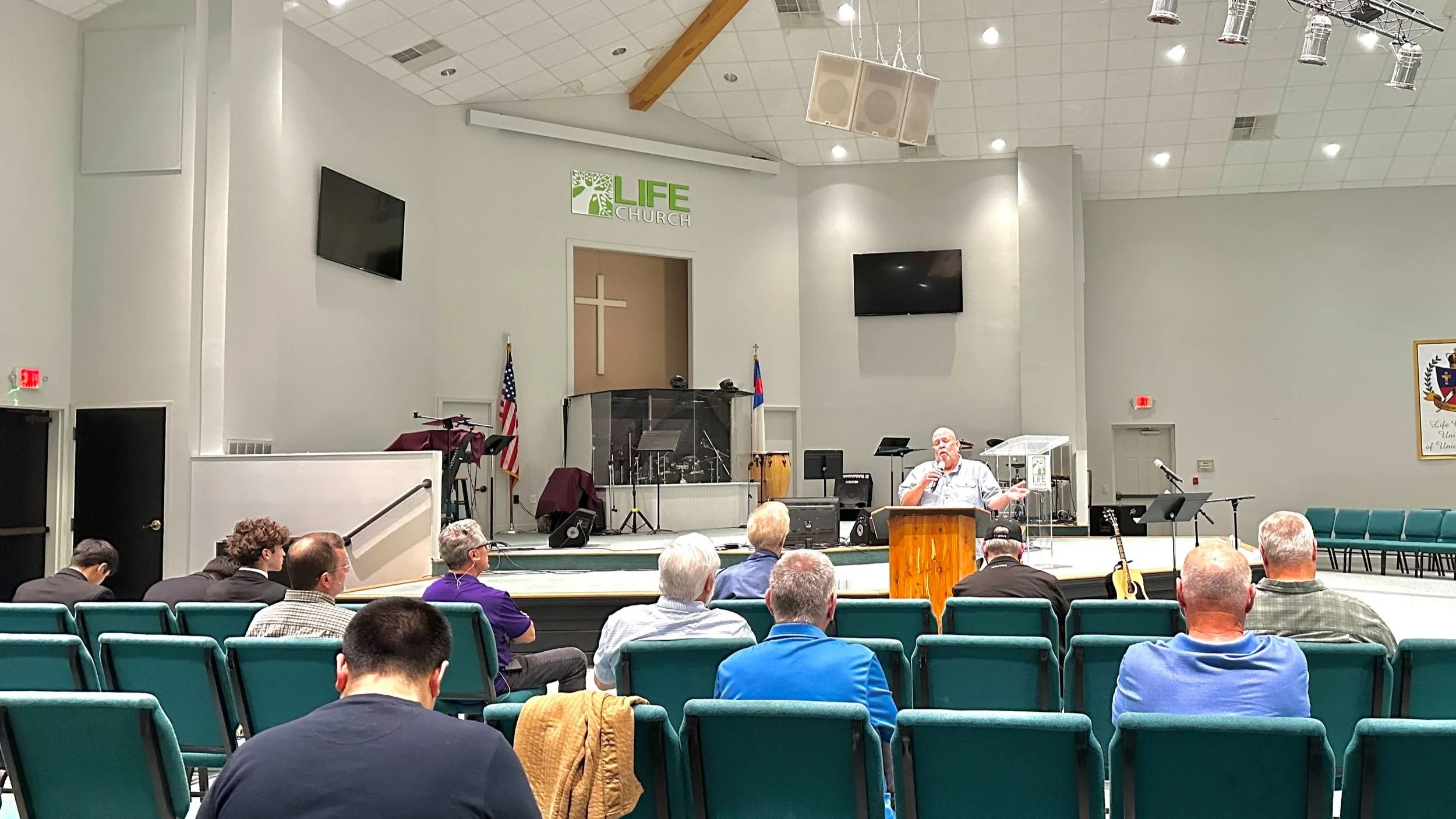 Interior of a church with a speaker at the pulpit addressing the congregation, rows of teal chairs, American flags, and musical instruments on stage. Men's Meeting