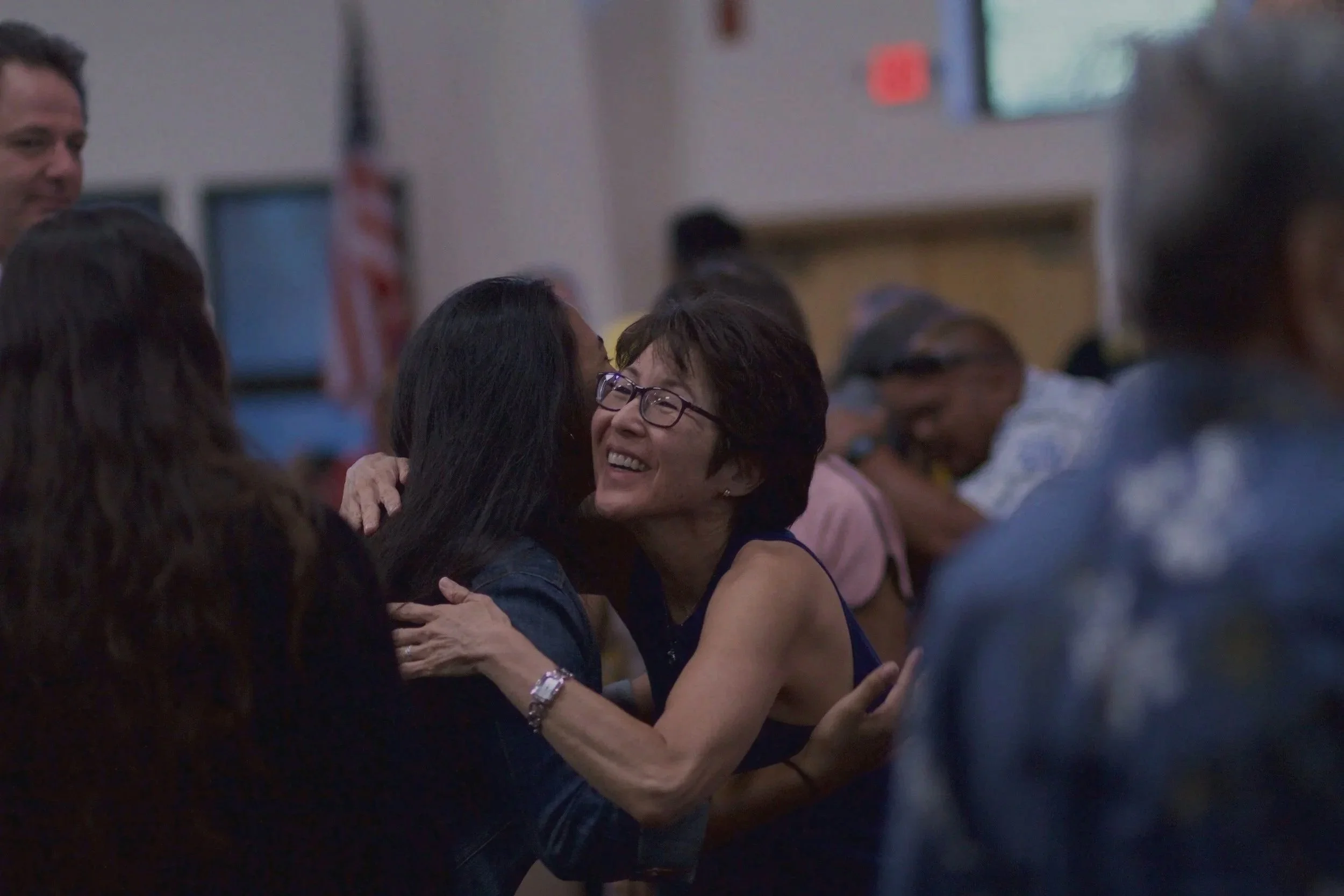 Two women hugging and smiling at each other in a crowded indoor setting.