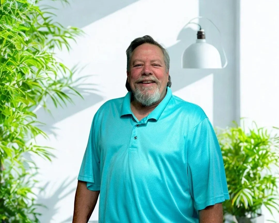 A smiling man with a beard, wearing a bright turquoise polo shirt, standing in front of a red semi-circle background on a gray wall.