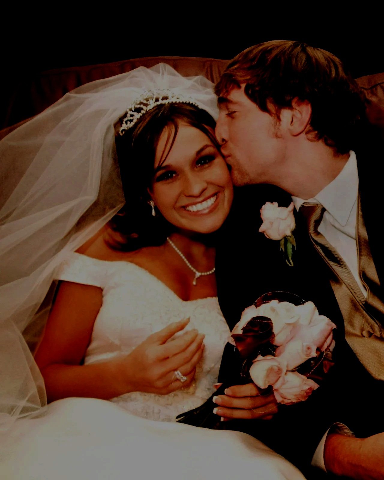 A bride and groom on their wedding day, with the groom kissing the bride's forehead. The bride is smiling and holding a bouquet of pink roses. The bride is wearing a white wedding dress, a pearl necklace, and a veil. The groom is dressed in a dark suit with a white shirt and a tie.