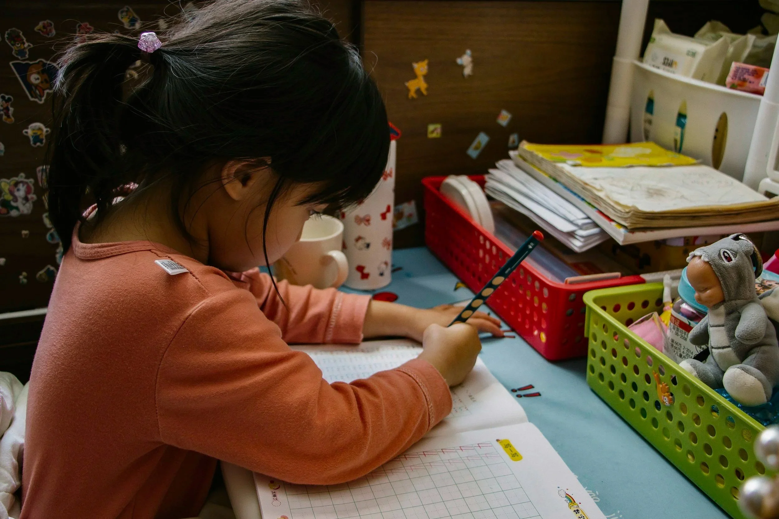A young girl with black hair tied in a ponytail, wearing a pink long-sleeved shirt, is sitting at a desk, writing in a notebook with a polka-dotted pencil.