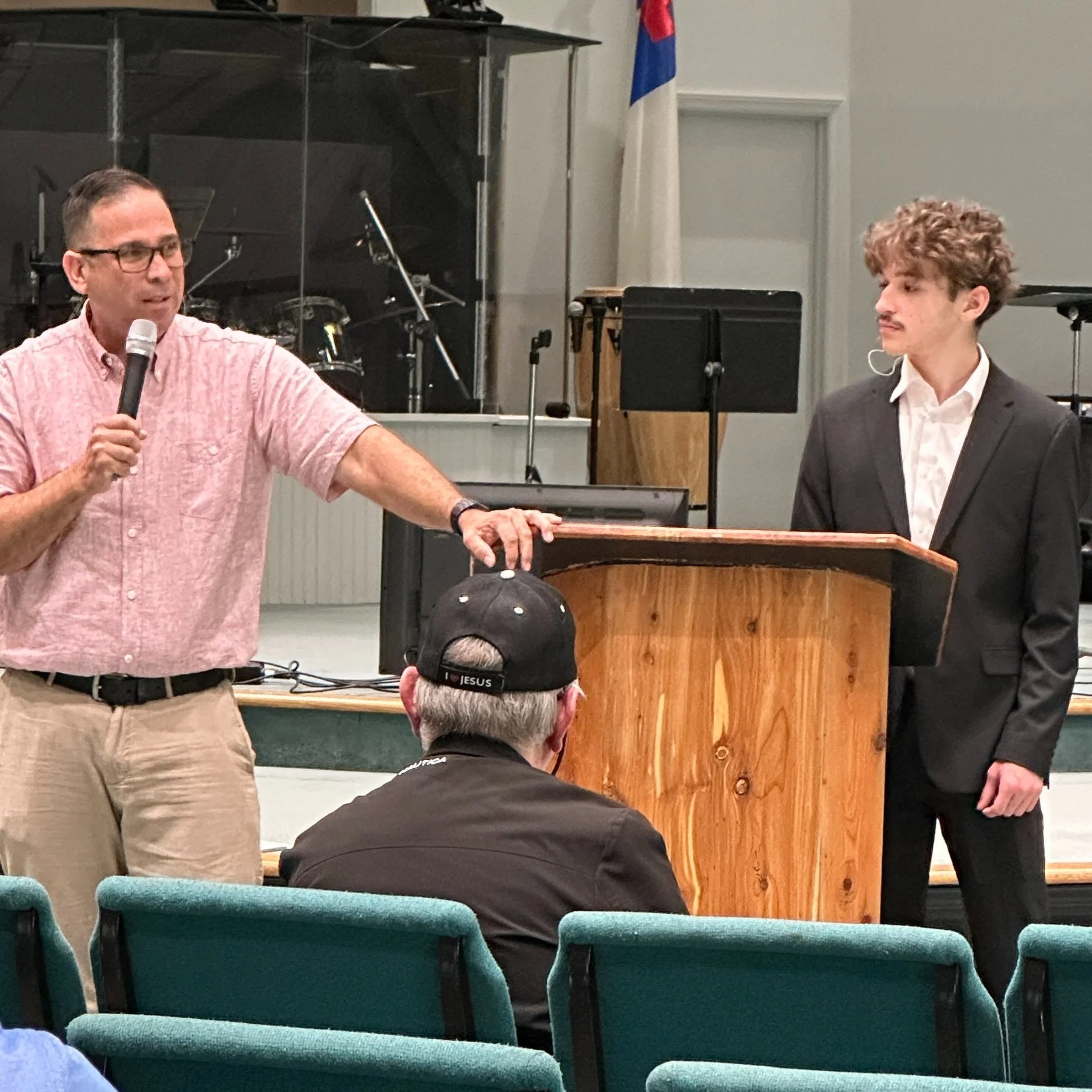 A man with glasses and a pink short-sleeve shirt speaking into a microphone, with his hand resting on a wooden podium. Next to him, a young man with curly blond hair wearing a black suit and white shirt stands. They are in a church or auditorium with green chairs, and an audience member with a black cap labeled 'JESUS' is seated in front of them.