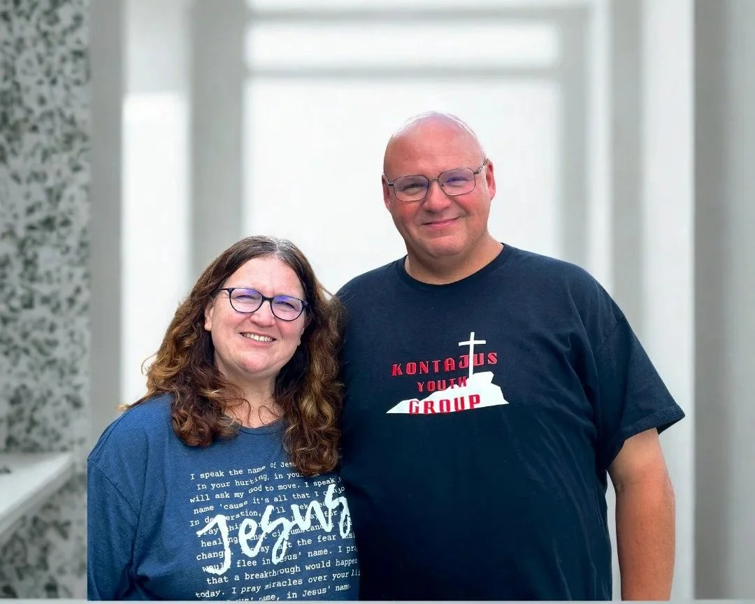 A smiling woman and man standing together against a red and gray background, both wearing T-shirts with religious and youth group slogans.