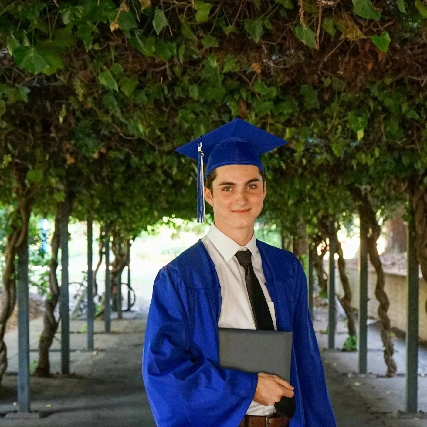 A young man in a blue graduation gown and cap holding a diploma, standing outdoors in a shaded area with green foliage and trees in the background.