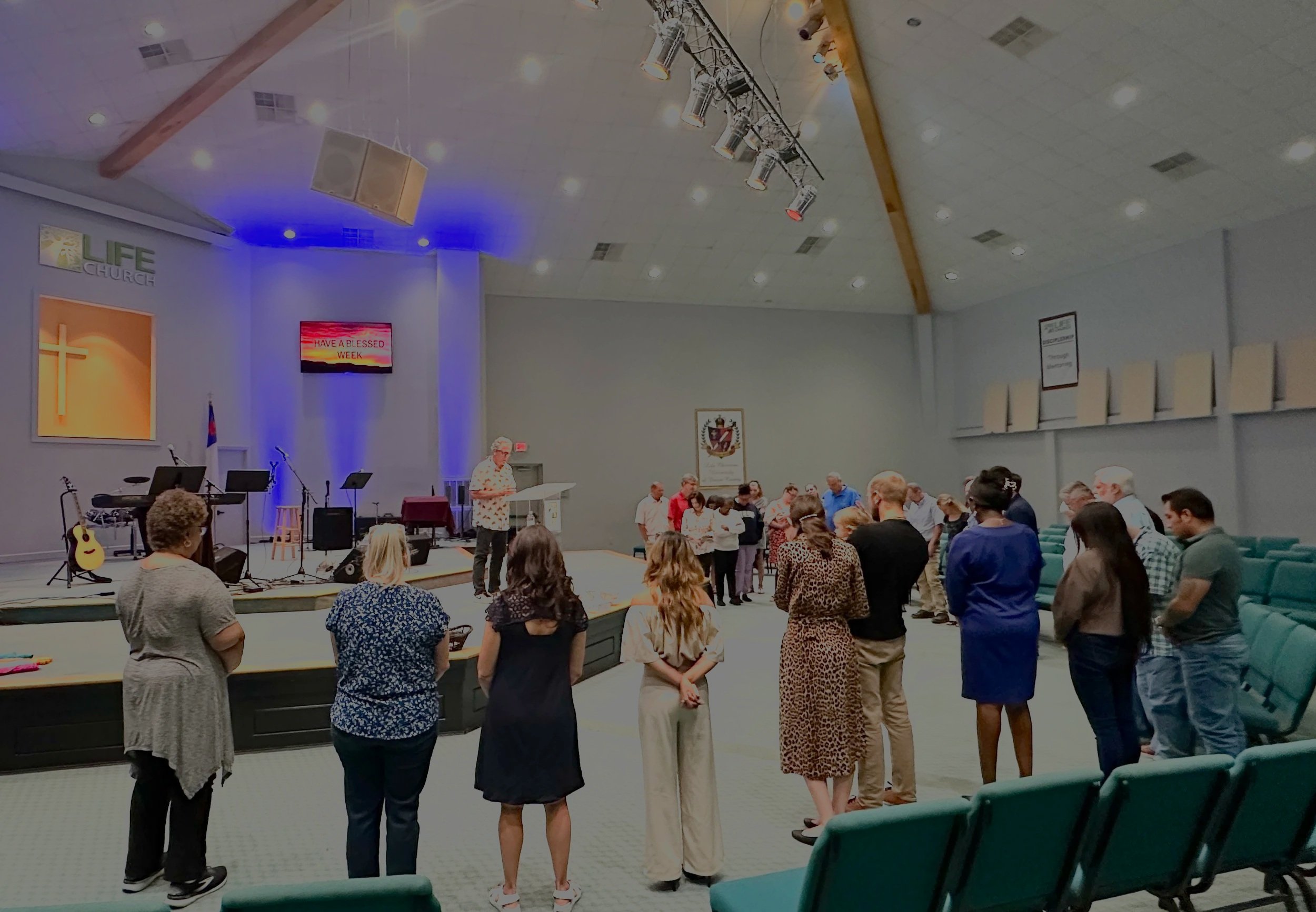 People praying and standing in a church auditorium during a worship service or prayer session. The stage has musical instruments and a large screen displaying the message "HAVE A BLESSED WEEK." The church interior includes a cross symbol and a flag on one side.
