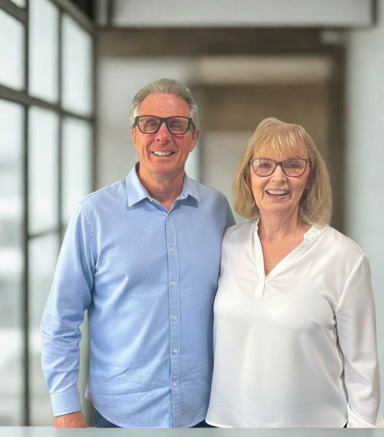 Smiling older man and woman standing together against a red circular backdrop, both wearing glasses and casual clothing.