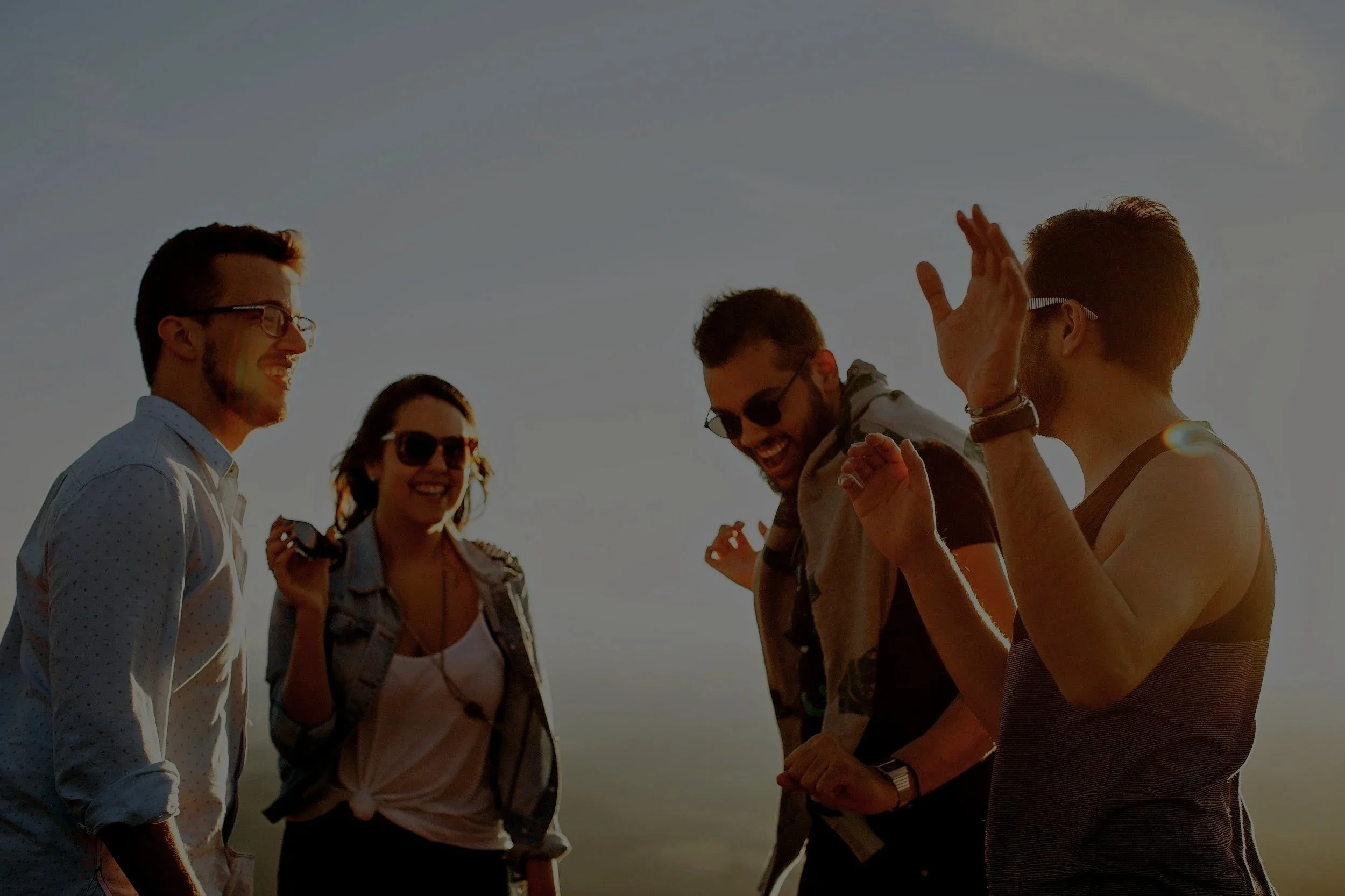 Four friends outdoors at sunset, laughing and chatting, with one holding sunglasses and another in a tank top, enjoying each other's company.