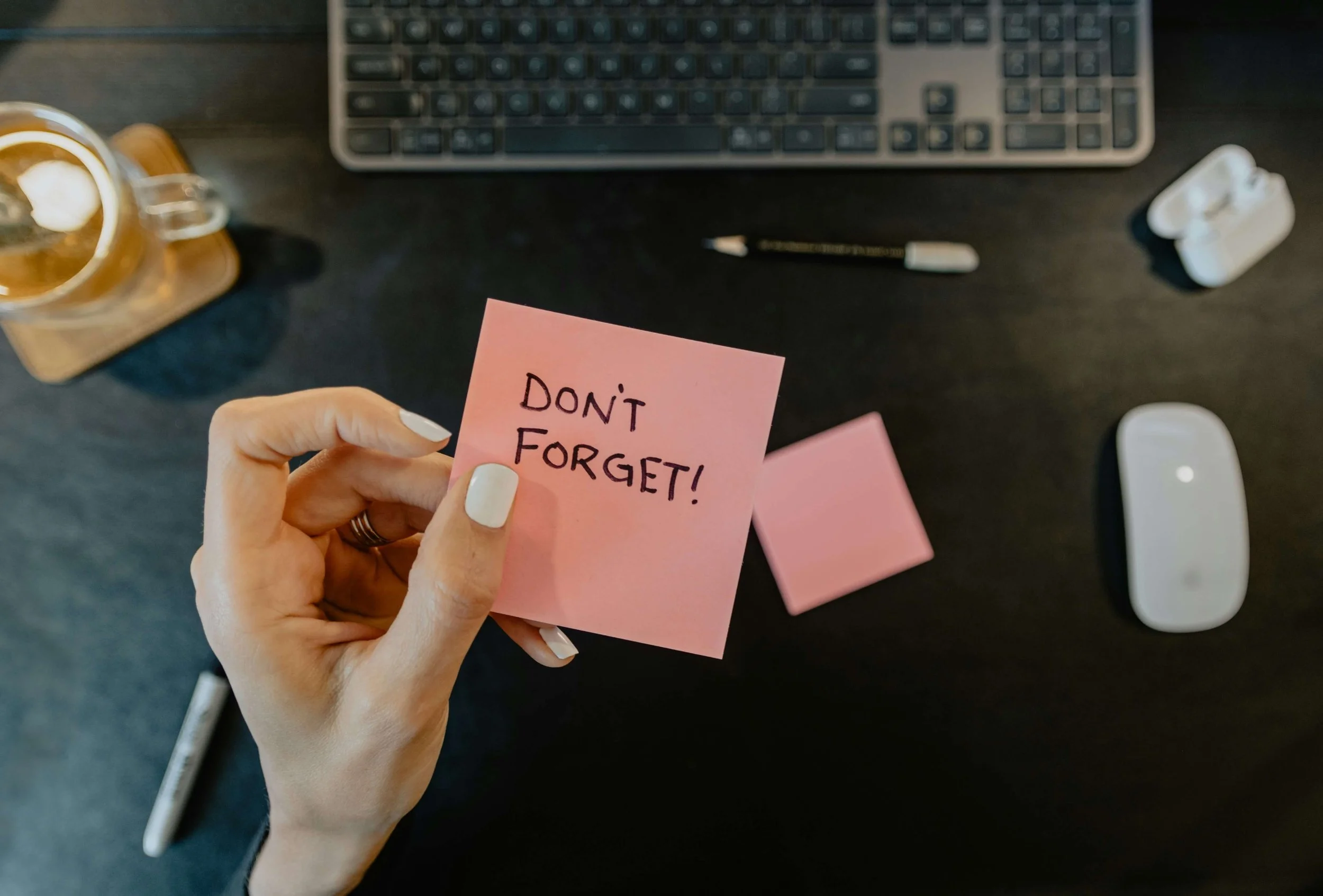 A white hand with white nail polish holding a pink sticky note that says 'Don't Forget.' The background is a desk with a keyboard and drink.