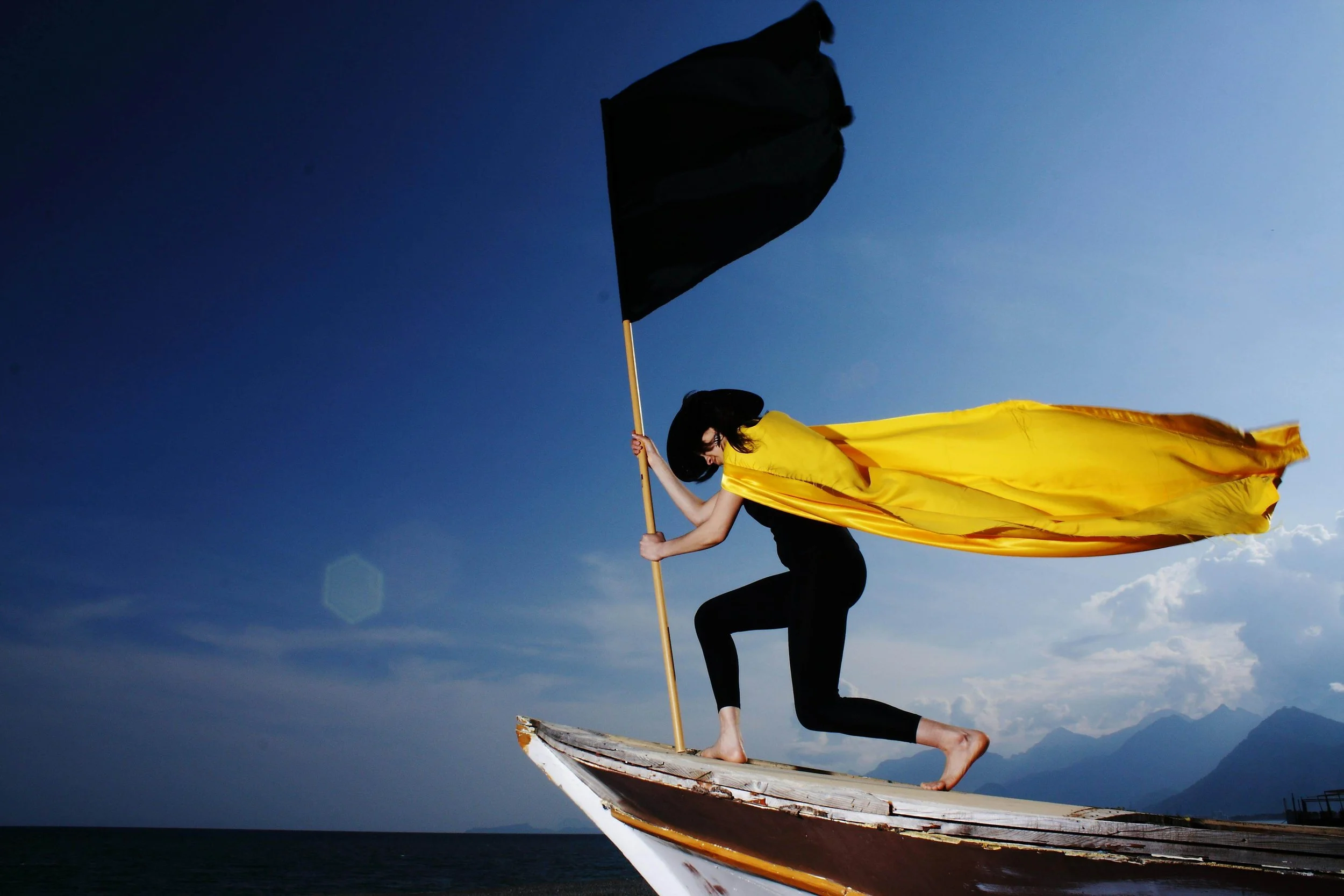 Woman on the front of a small boat holding a black flat, wearing a black hat and yellow cape. Represents perfectionist tendencies and anxiety therapy in Seattle during your fertility journey.