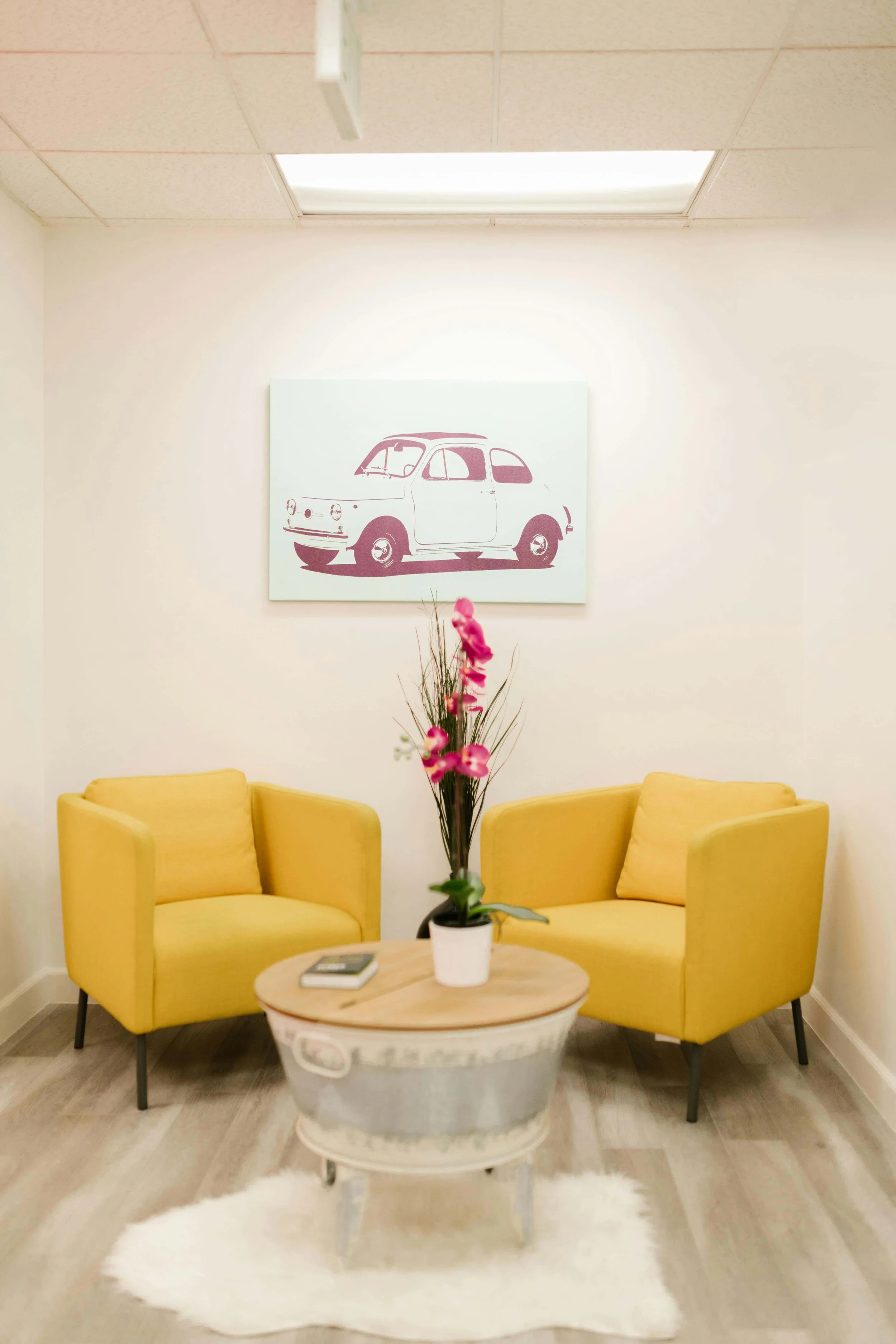 A waiting room with two yellow chairs, a small round table with pink flowers and a book on it. An art piece on the wall of a small car. Represents medical trauma therapy Seattle