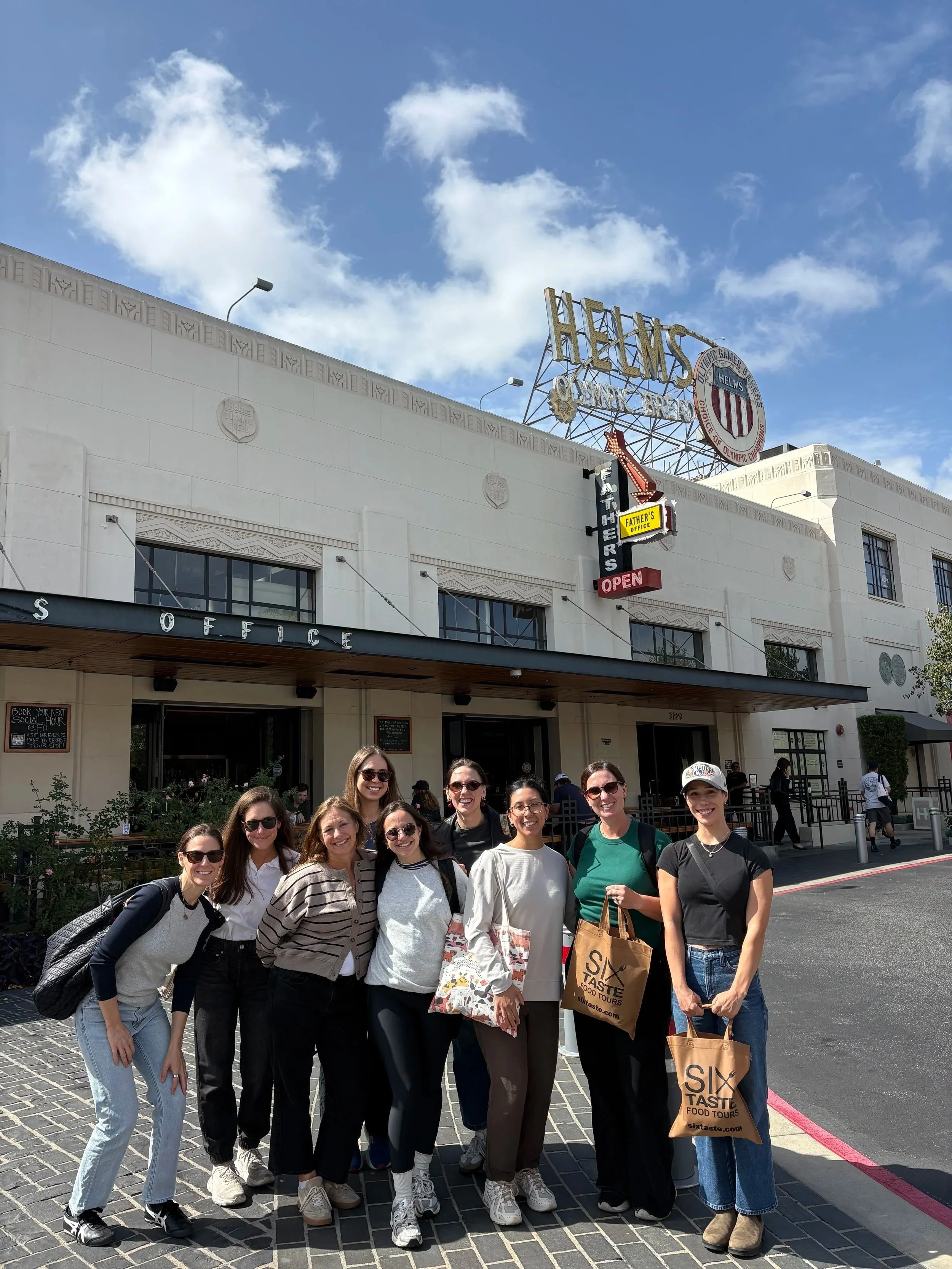 Group of smiling women standing outside in front of a building with a sign that reads 'HELEN'S' and a logo with a shield, along with other signs indicating food tours, under partly cloudy sky.