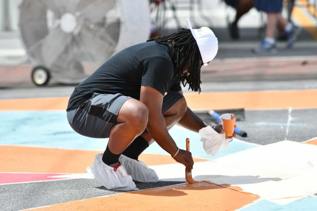 Person crouching on the ground painting a colorful mural, wearing a white cap, protective shoe covers, and black shorts.