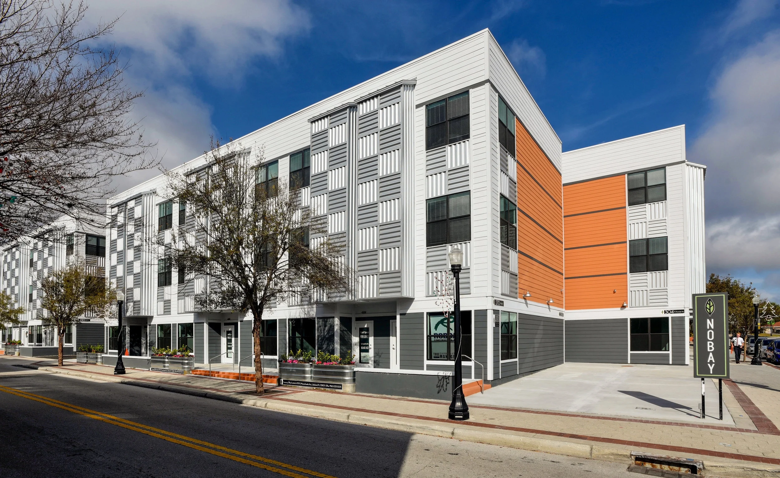 Modern multi-story apartment building with white, gray, and orange exterior siding, sidewalk with trees, street lamps, and a sign that reads 'NOBAY' under a partly cloudy blue sky.