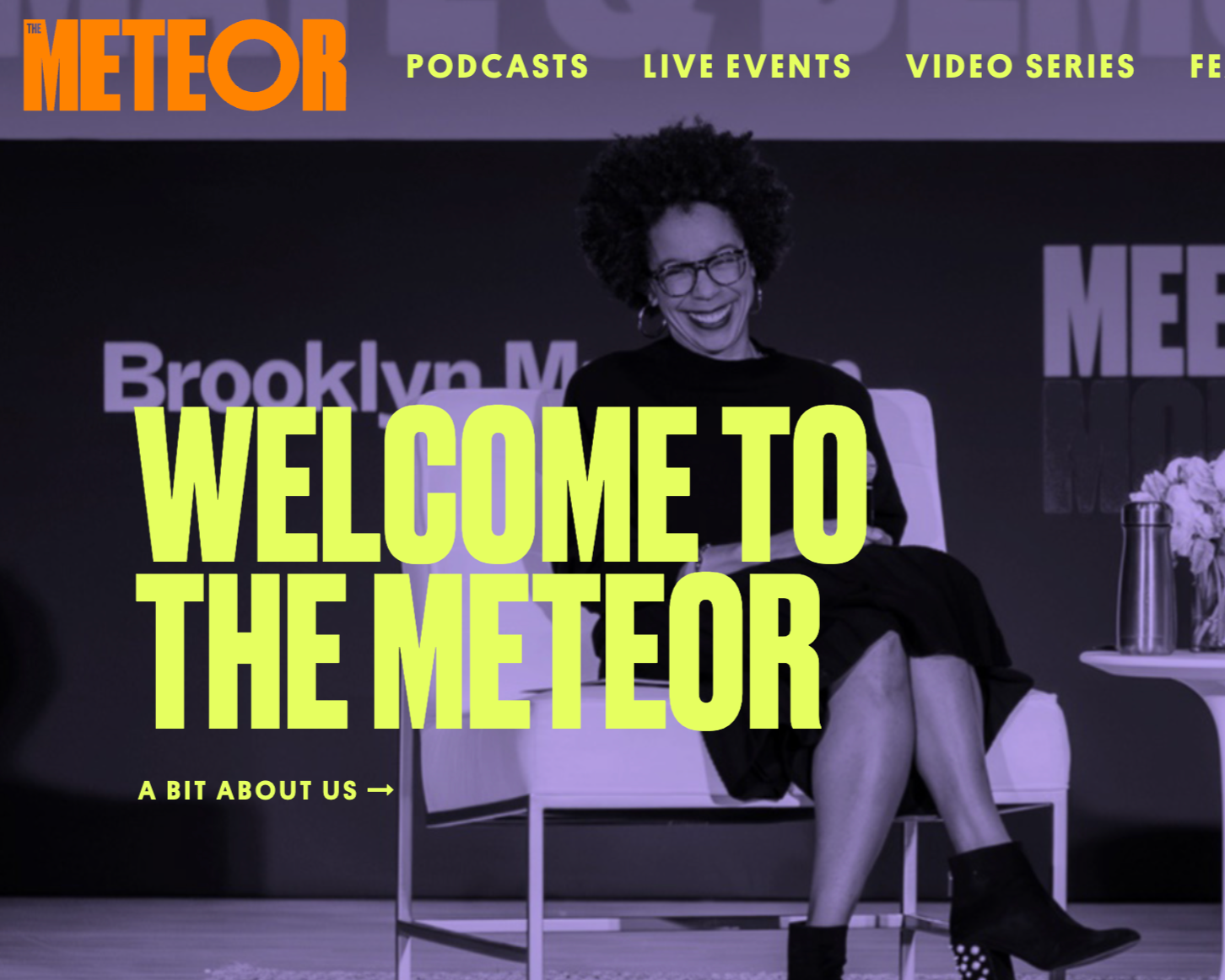 A woman sitting on a white chair, smiling and wearing glasses, at an event with a dark background and brightly colored text that reads 'Welcome to The Meteor'.