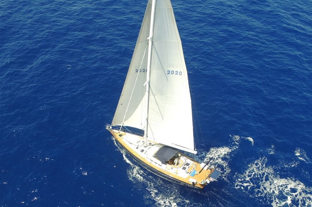    Aerial view of a sailboat gliding on a vast blue ocean, its white sails fully extended displaying the numbers "2020 0505". The boat leaves a gentle trail of white foam in its wake. The deep blue water surrounds the boat, providing a stark contrast