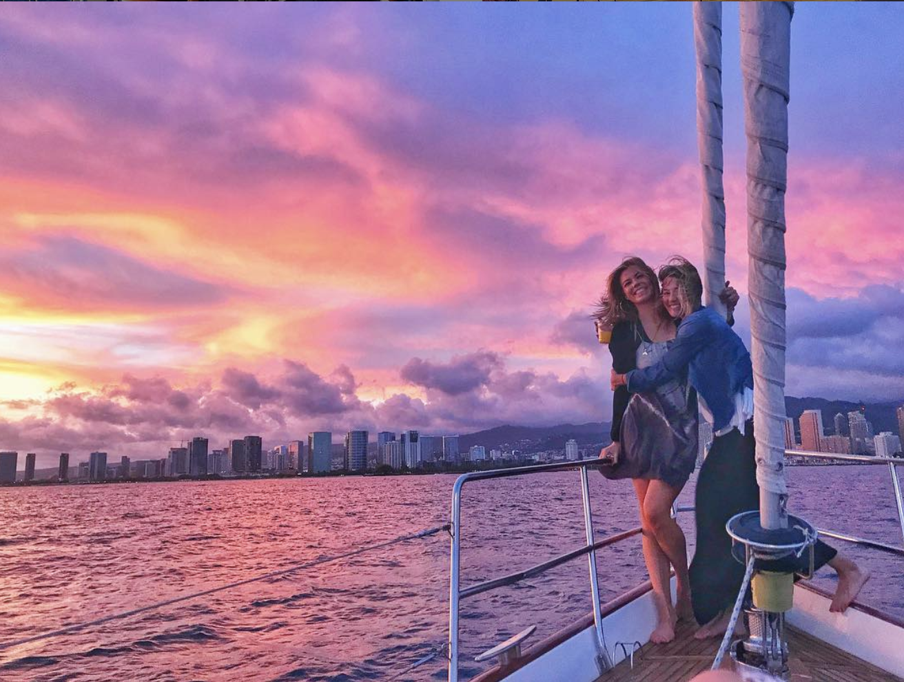    Two individuals sharing a joyful moment aboard a sailboat during a breathtaking sunset. The sky is painted in hues of pink, purple, and orange, casting a warm glow over the city skyline in the distance. The duo stands close, one holding a drink, w