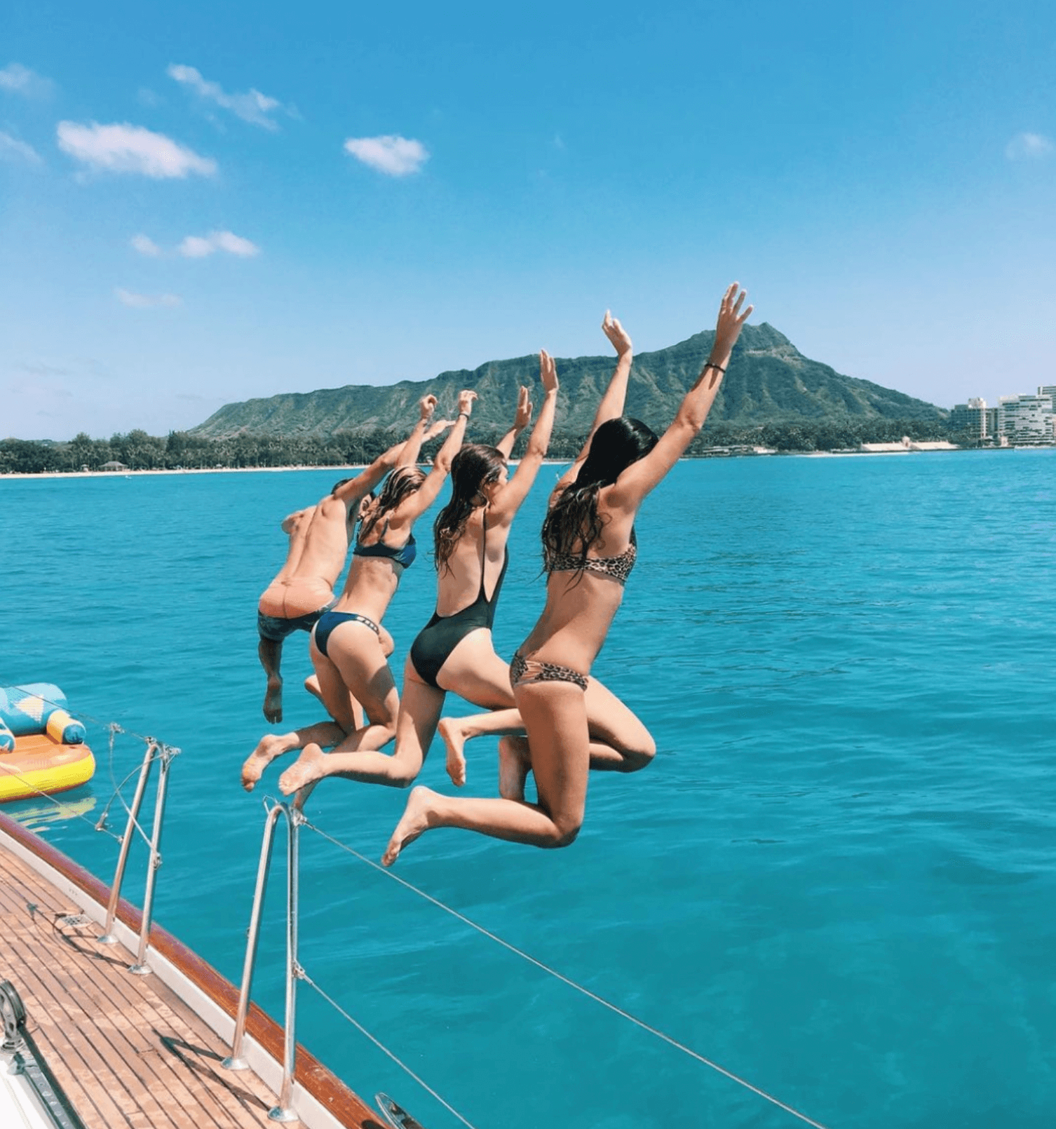    Four individuals joyfully leaping off the side of a sailboat into the crystal-clear turquoise waters, arms raised in excitement. Their poised mid-air poses capture the essence of adventure and freedom. In the background, a scenic coastline with lu