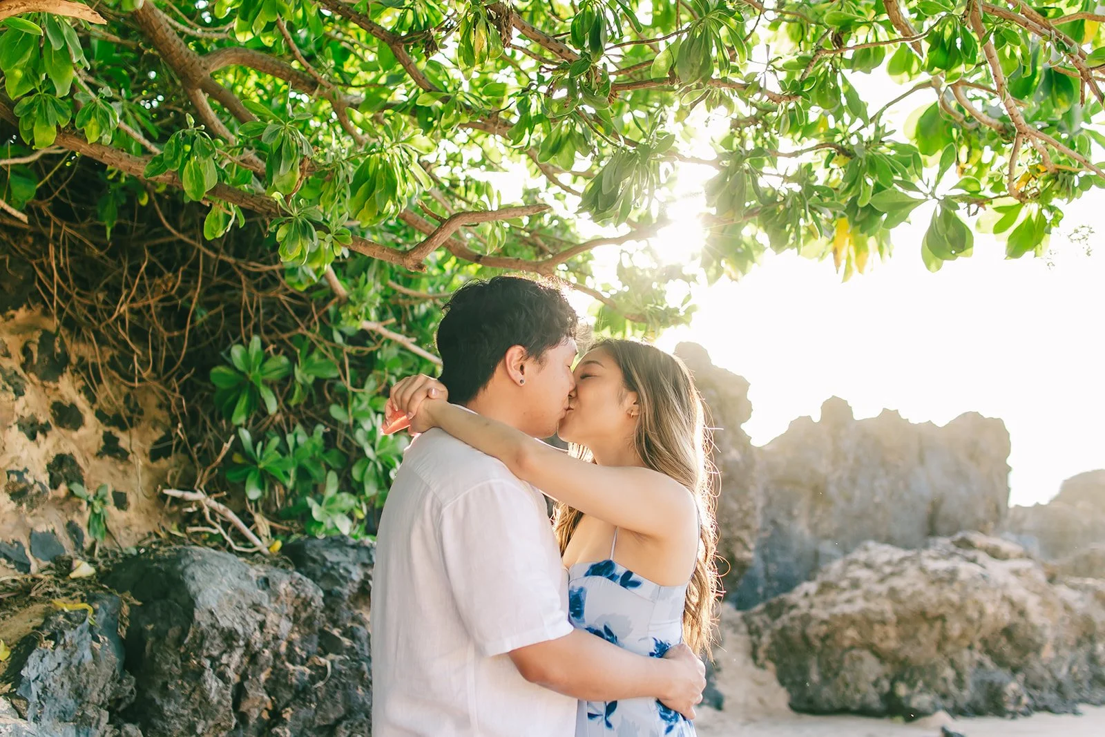  An Asian couple in Hawai’i with the most beautiful and bright lighting that makes the photo so pretty! They are both kissing each other, and the woman’s hand is on his shoulder and neck, while the man is holding her waist. They are sharing a close c
