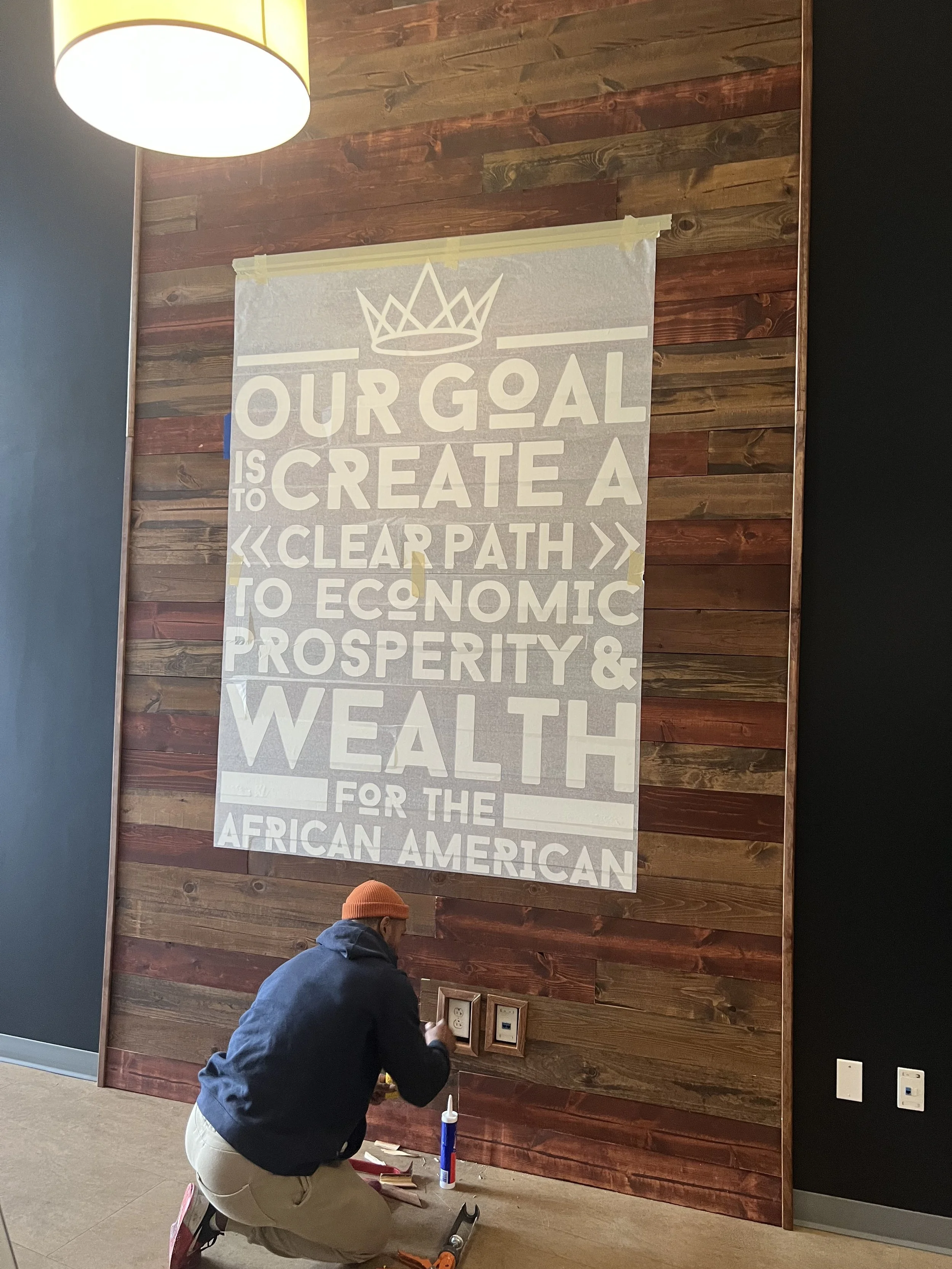 KC G.I.F.T. statement wall installation showing typography graphic being applied over reclaimed wood, bringing the organization’s mission into the space.
