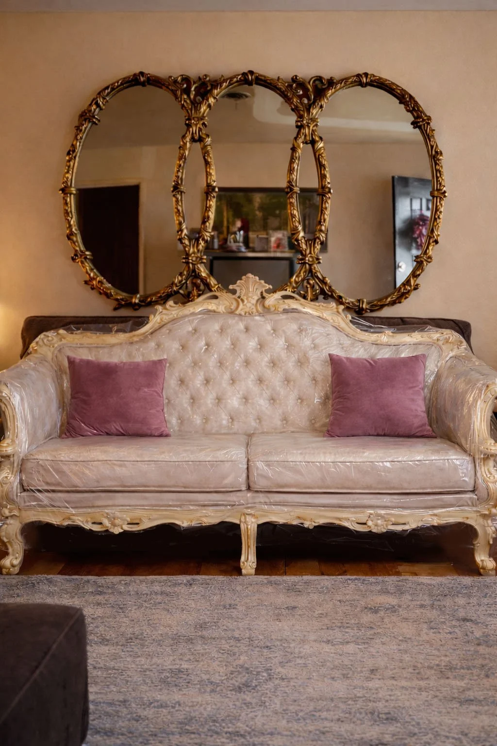 Vintage cream Rococo sofa wrapped in clear protective plastic, styled with two mauve velvet pillows and placed beneath ornate gold mirrors in a curated, design-forward living room.