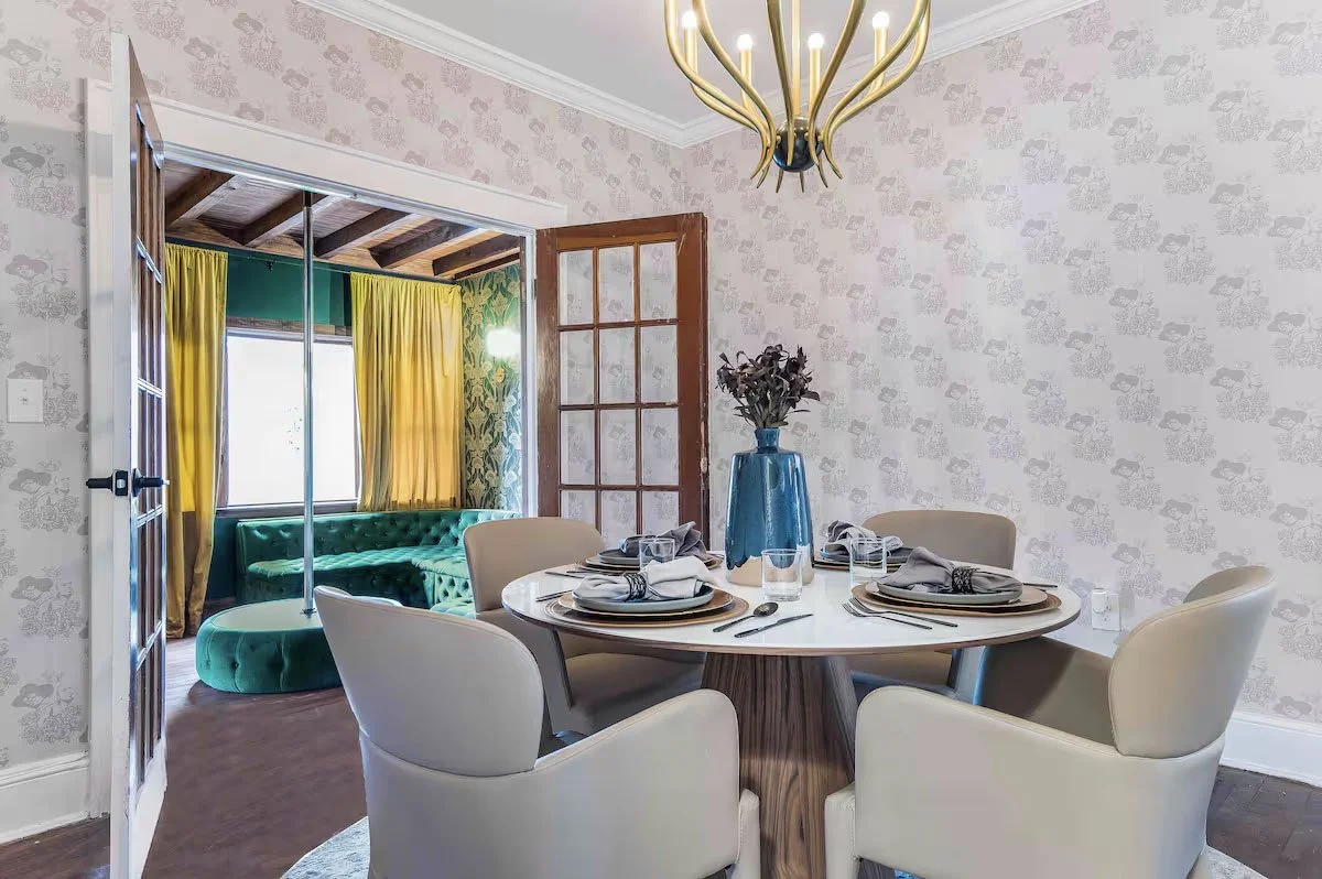 Dining room with custom illustrated wallpaper, round table, cream chairs, gold chandelier, and view into an adjoining green lounge.
