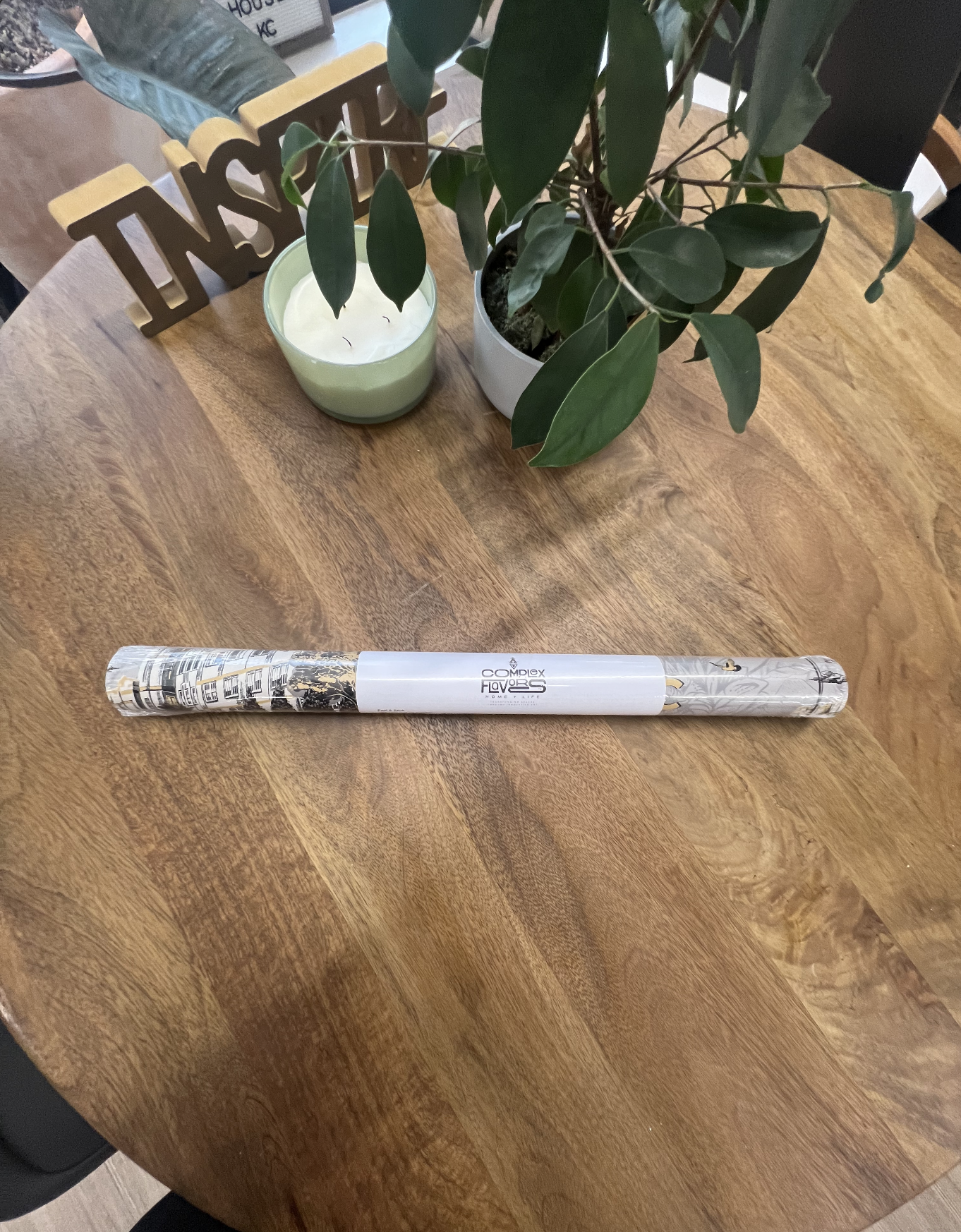 Branded sample roll of the custom Porter House KC toile wallpaper displayed on a wooden table beside plants and décor.