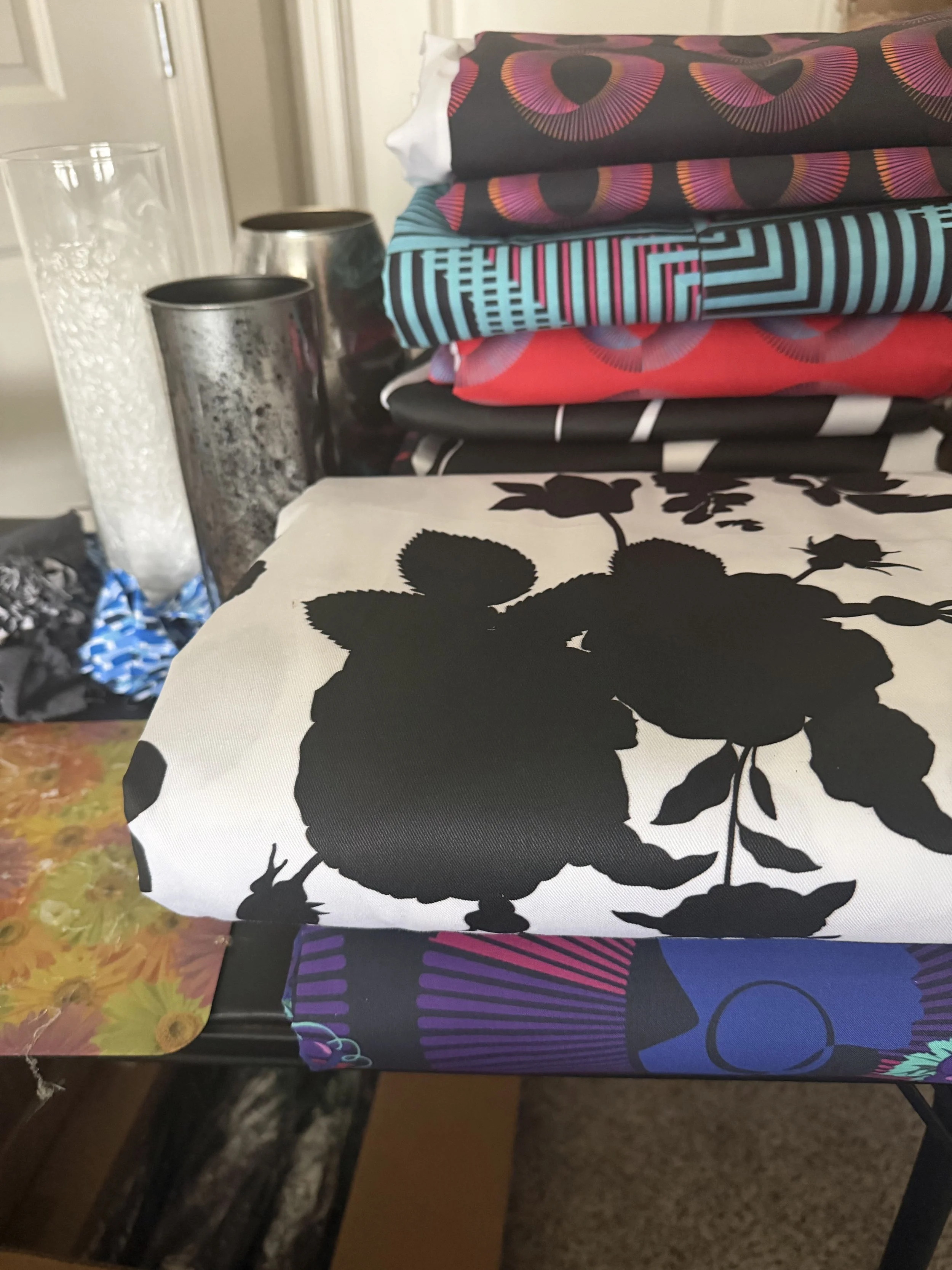 Stack of colorful patterned fabrics beside metallic and glass vessels during the design process.
