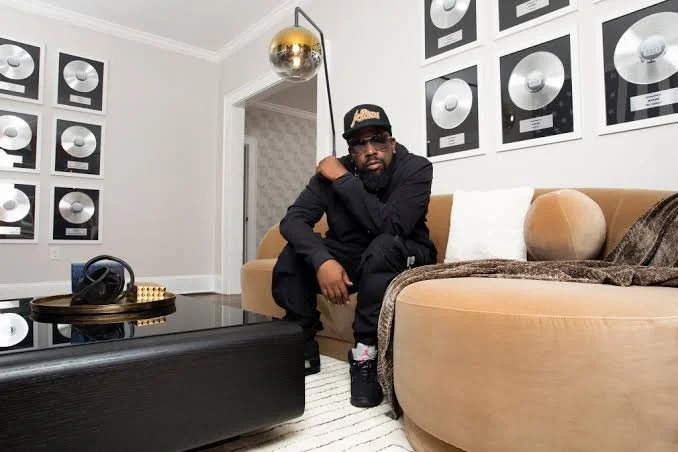 Big Boi seated in a living room surrounded by framed record plaques, neutral furniture, and modern decor.