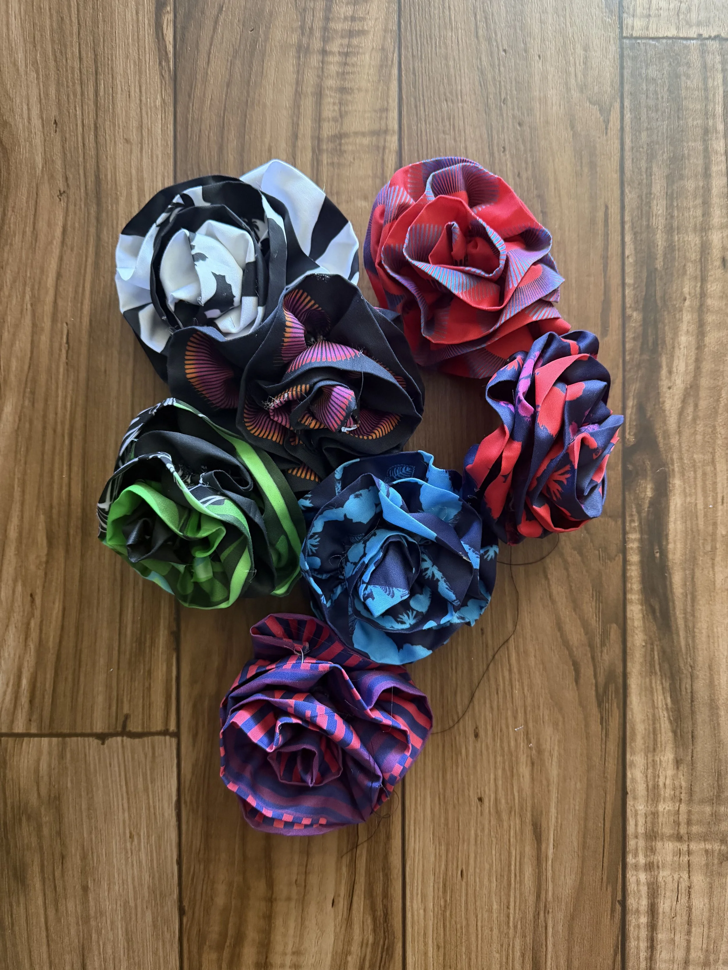 Seven handmade fabric flowers in colorful graphic patterns arranged on a wood floor.