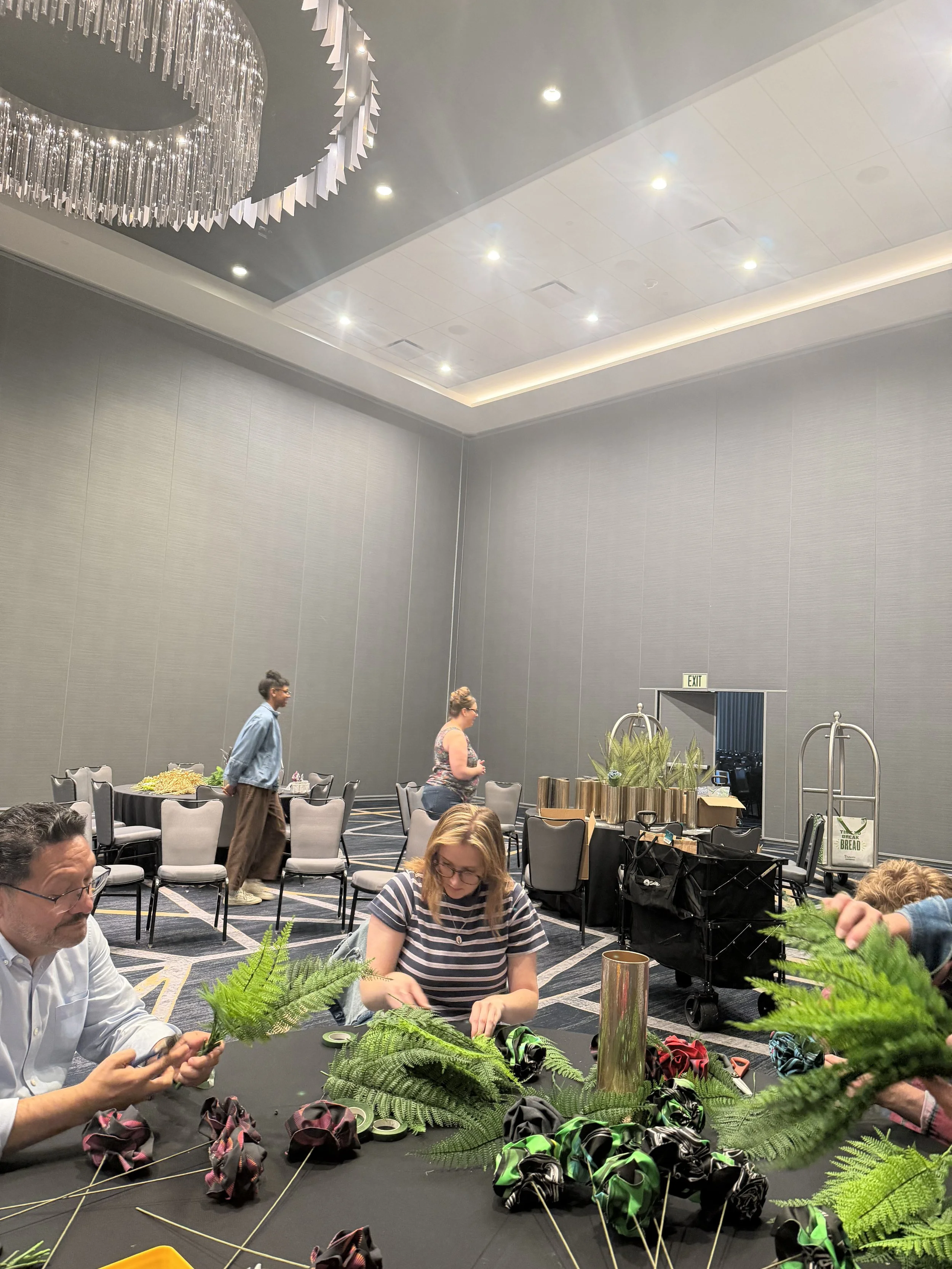 People assembling colorful fabric floral centerpieces with green fern foliage at an event venue.