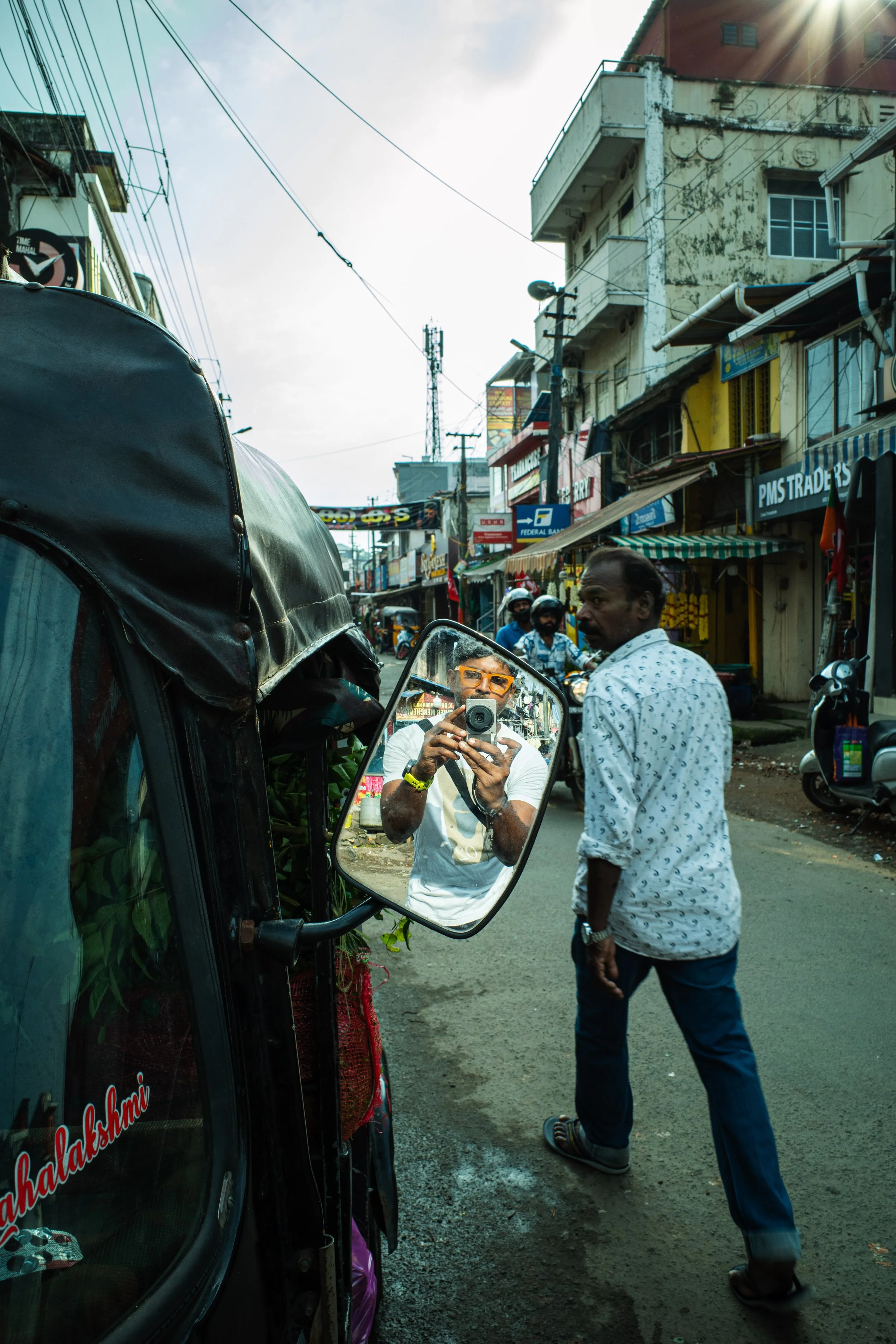 A man with orange sunglasses takes a photo of himself in a vehicle's side mirror that reflects the street scene, which includes a man walking and shops on a busy street with motorcycles and utility poles.
